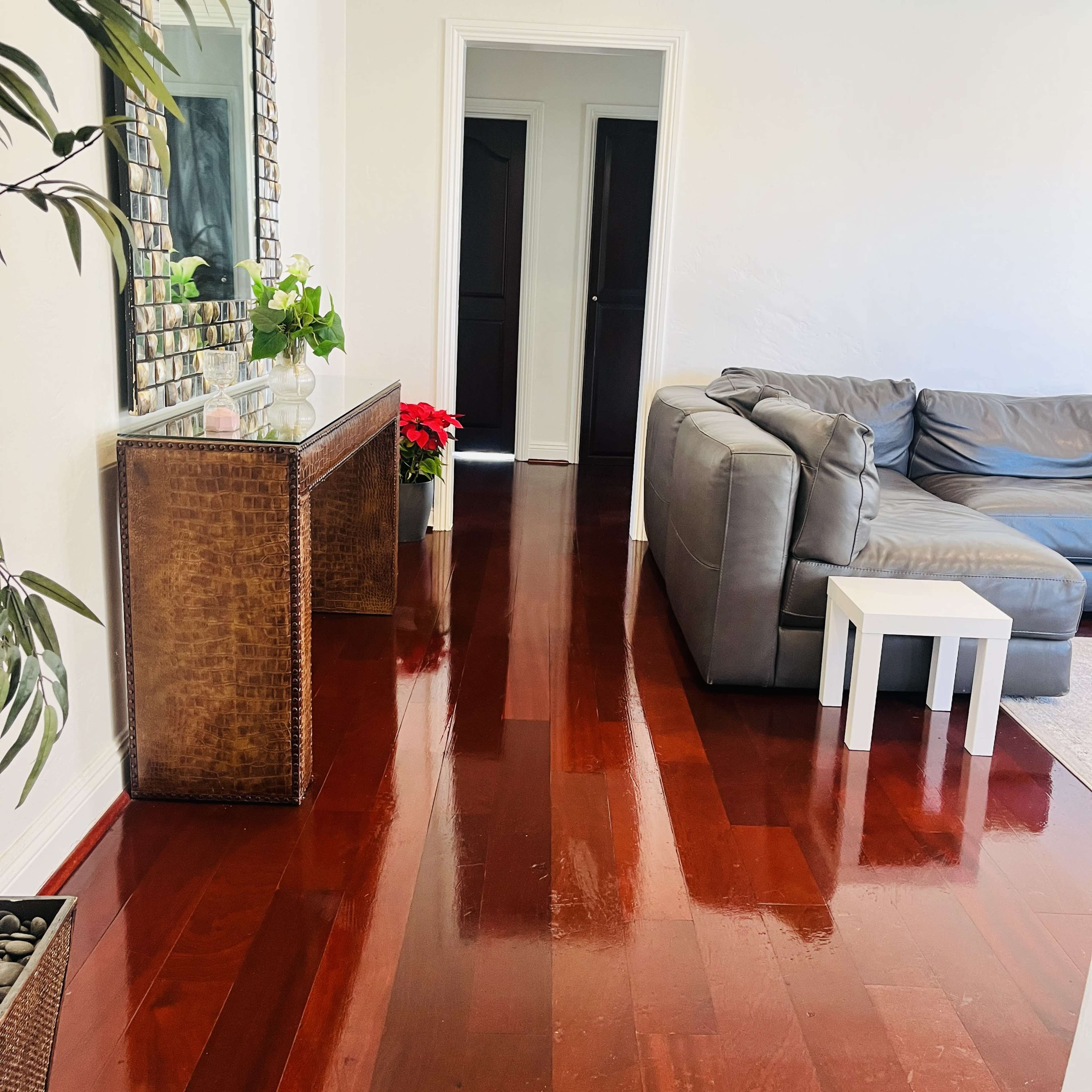 A living room with polished wooden floors, a gray sectional sofa, a glass-top console table with plants, and a hallway leading to a closed door.