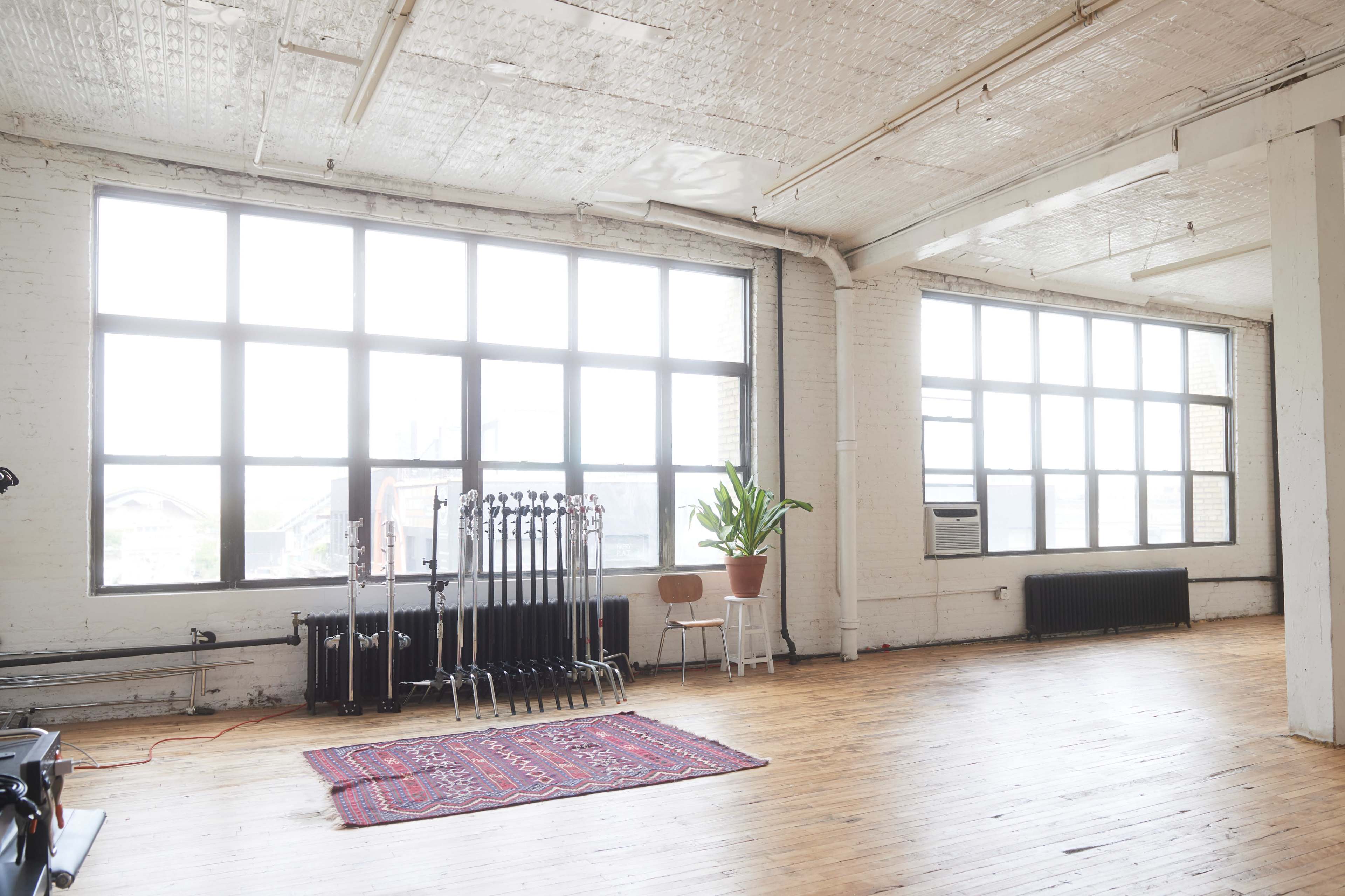 A spacious, industrial-style room with large windows, wooden floors, and a few chairs arranged alongside a potted plant.
