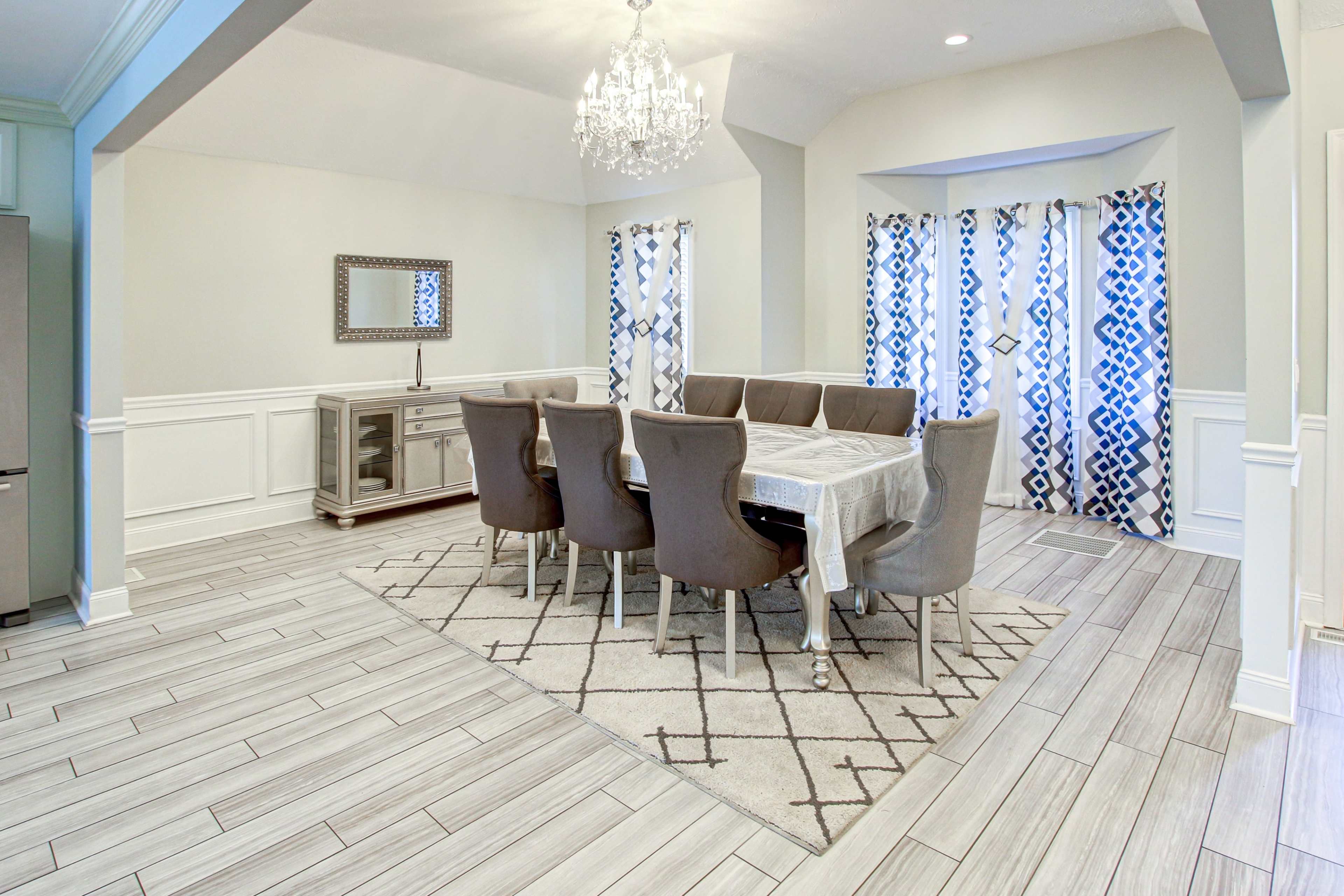 A modern dining room features a large table surrounded by upholstered chairs, a decorative chandelier above, and large windows with patterned curtains.