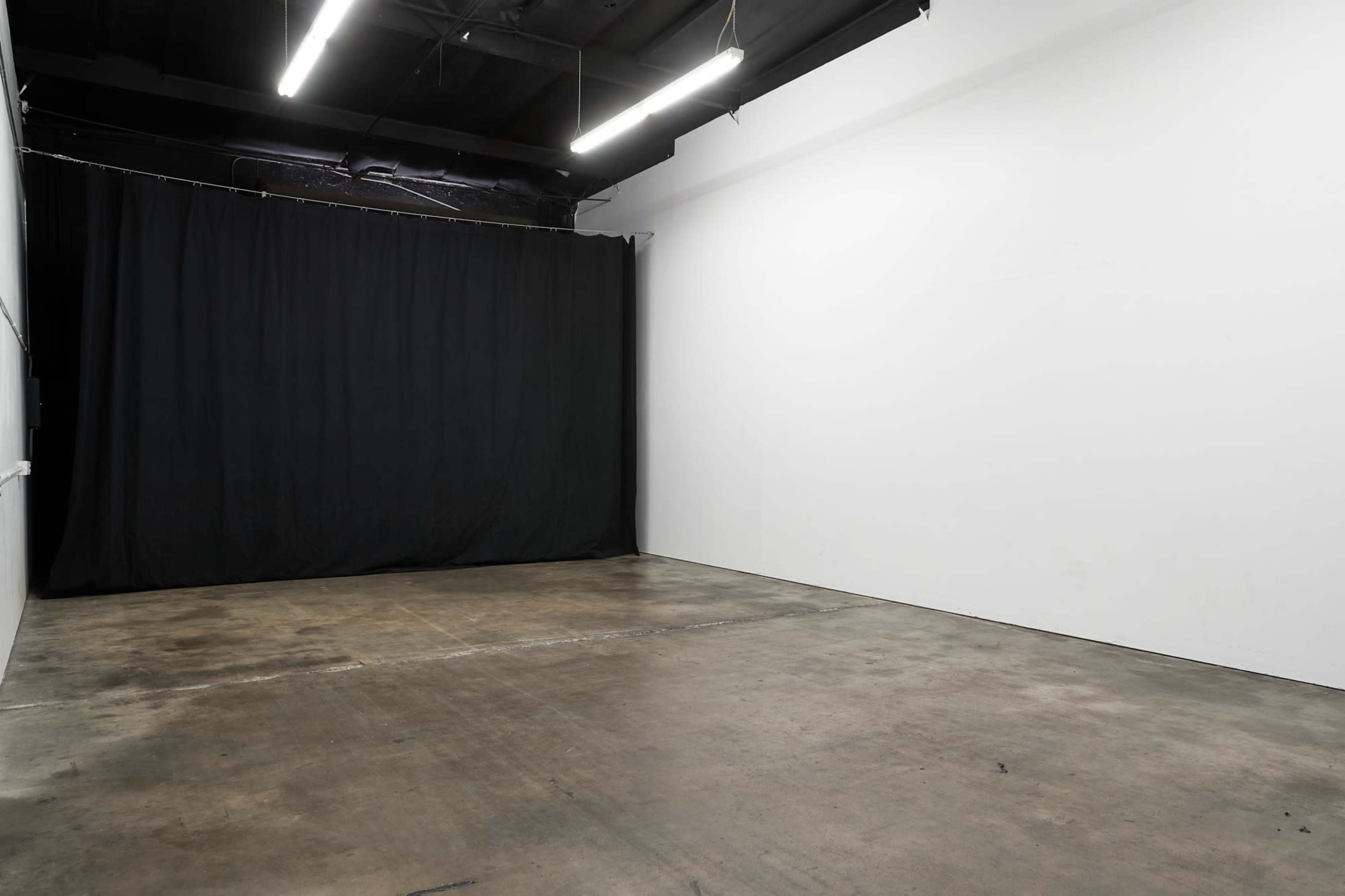 A stark, empty room with a concrete floor, featuring a black curtain along one wall and a plain white wall opposite it.