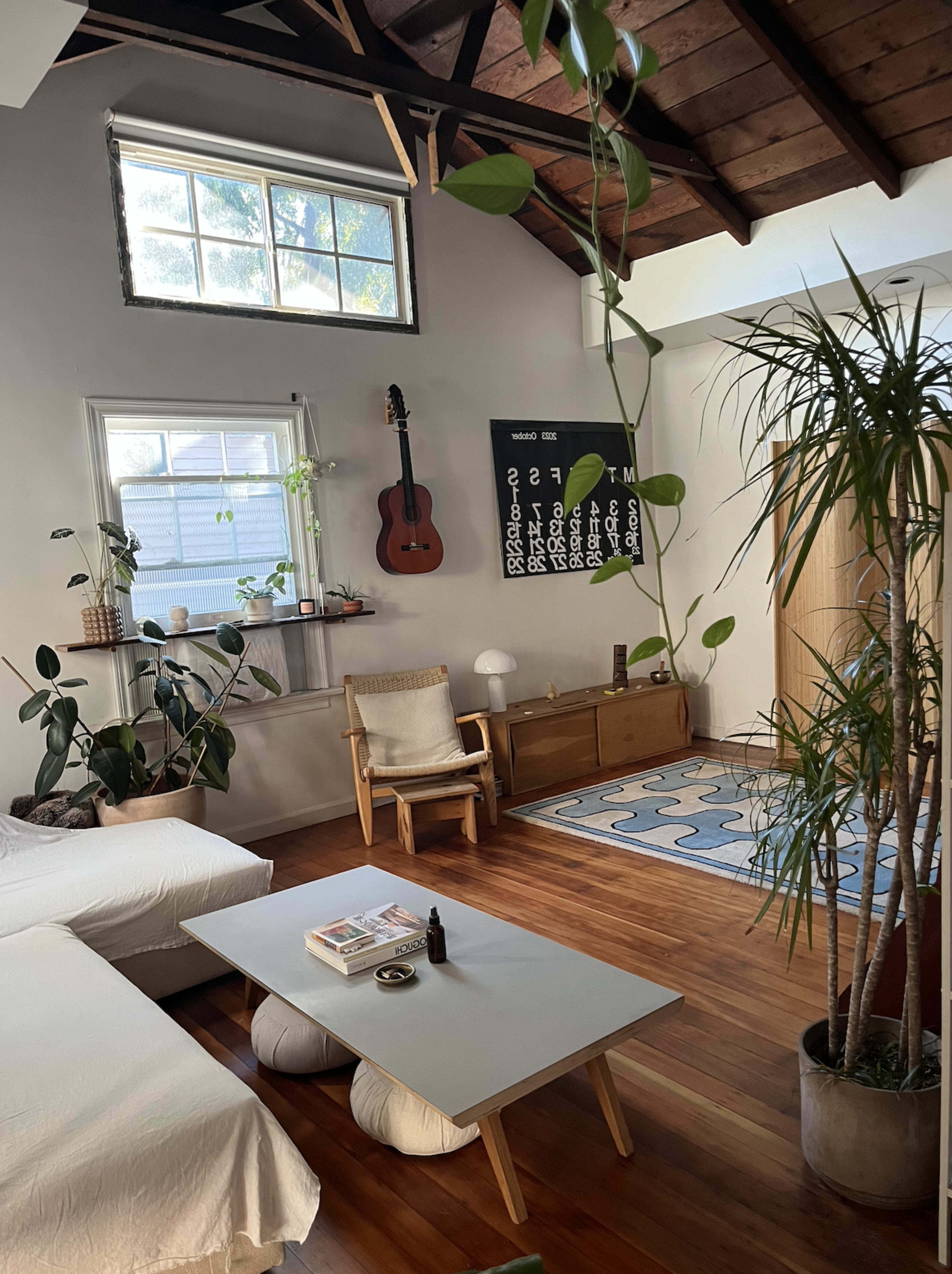 A cozy living room features a large window, a wooden ceiling, a guitar on the wall, and indoor plants surrounding a low coffee table and two poufs.