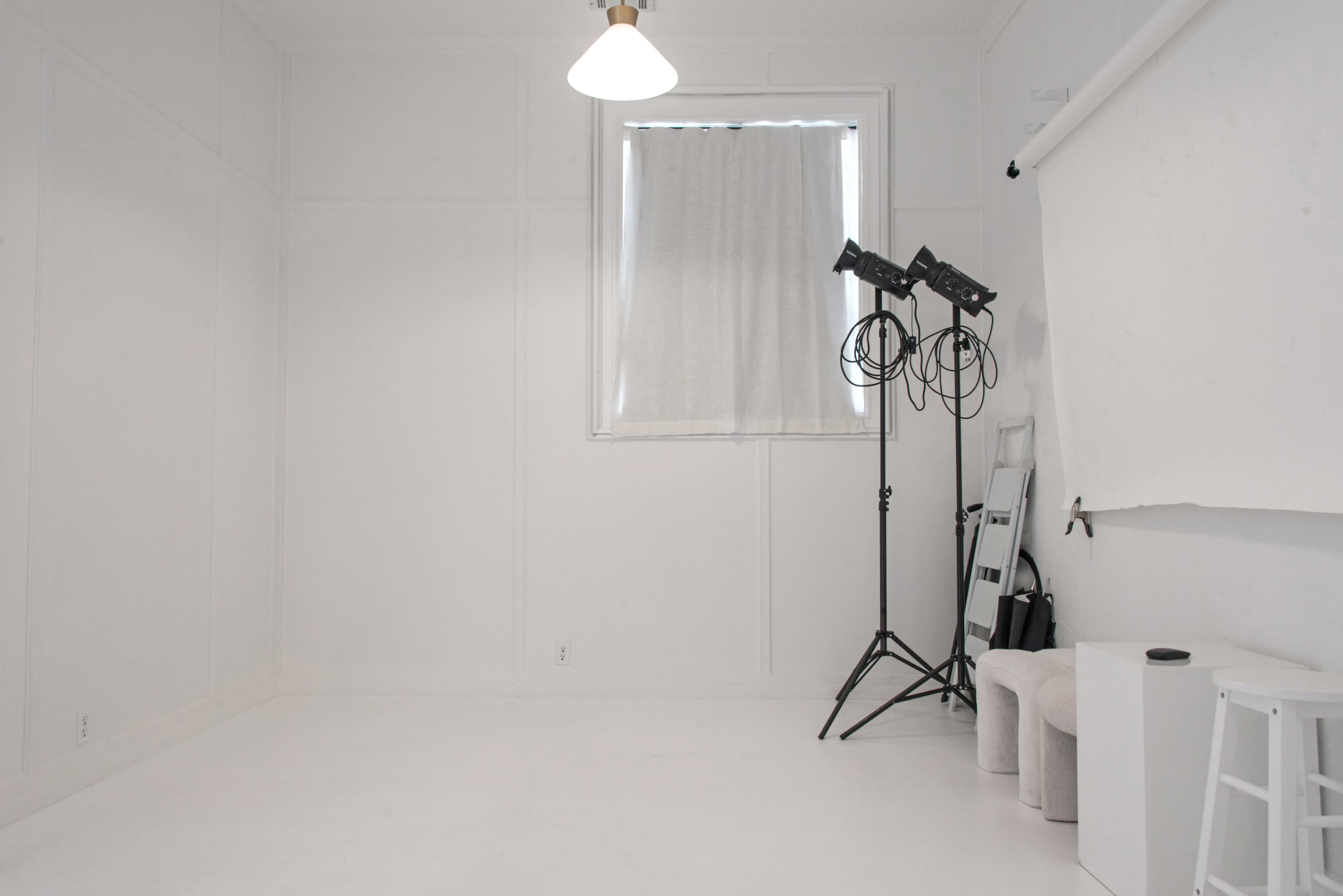 The image shows a sparsely furnished, white-walled room with a lighting setup, a chair, and a mirror.