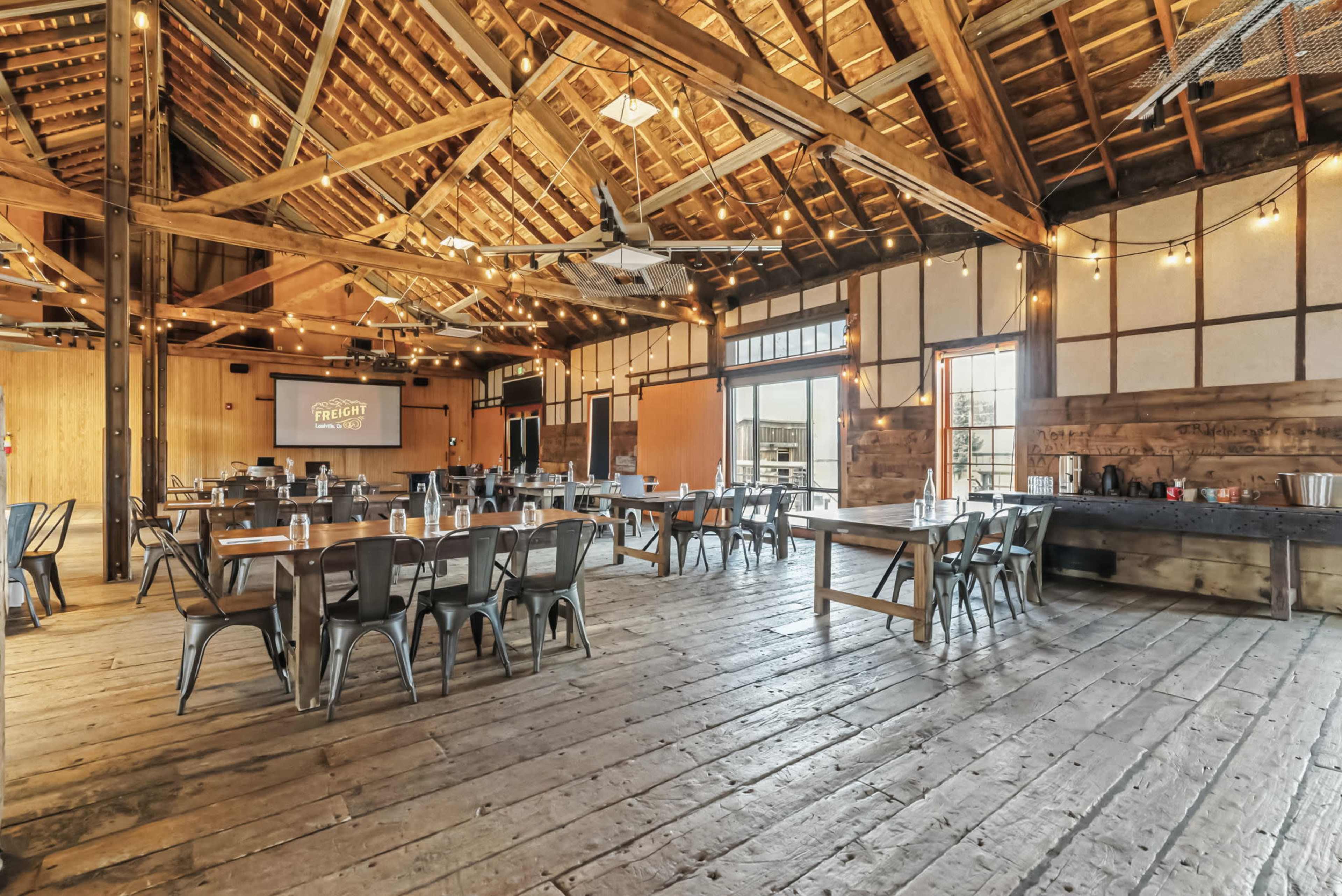 The interior of a spacious rustic event venue features wooden beams and a polished wood floor, with multiple dining tables arranged for a gathering.
