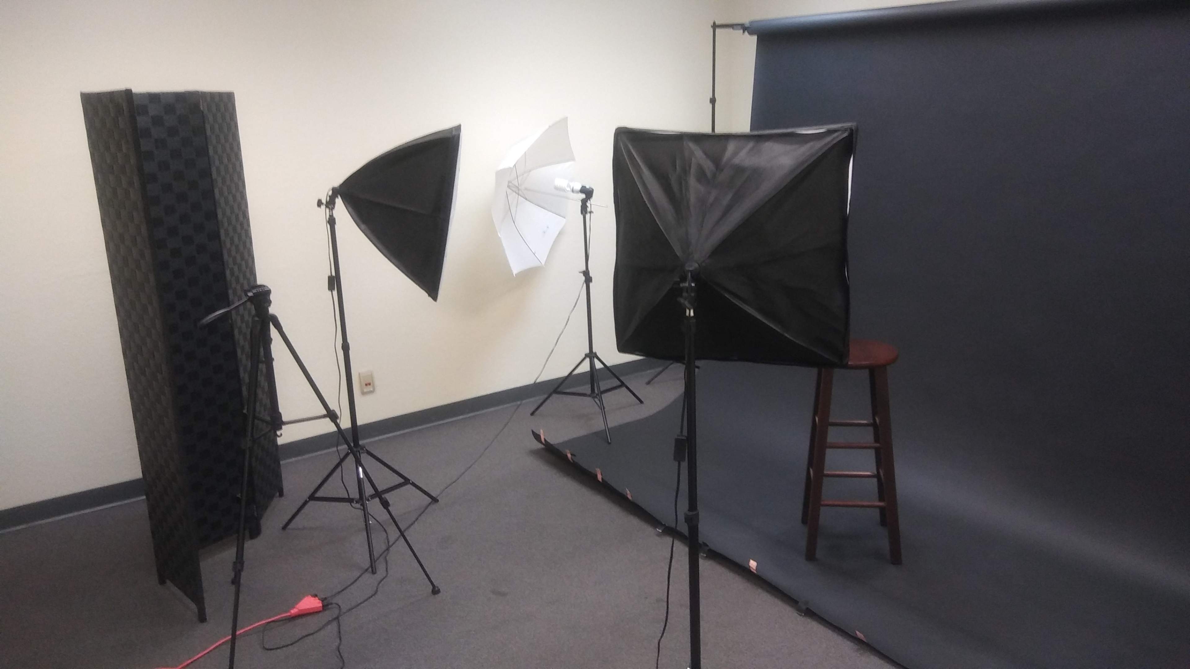 A photography studio setup with three lighting softboxes, a microphone stand, and a wooden stool in front of a dark backdrop.