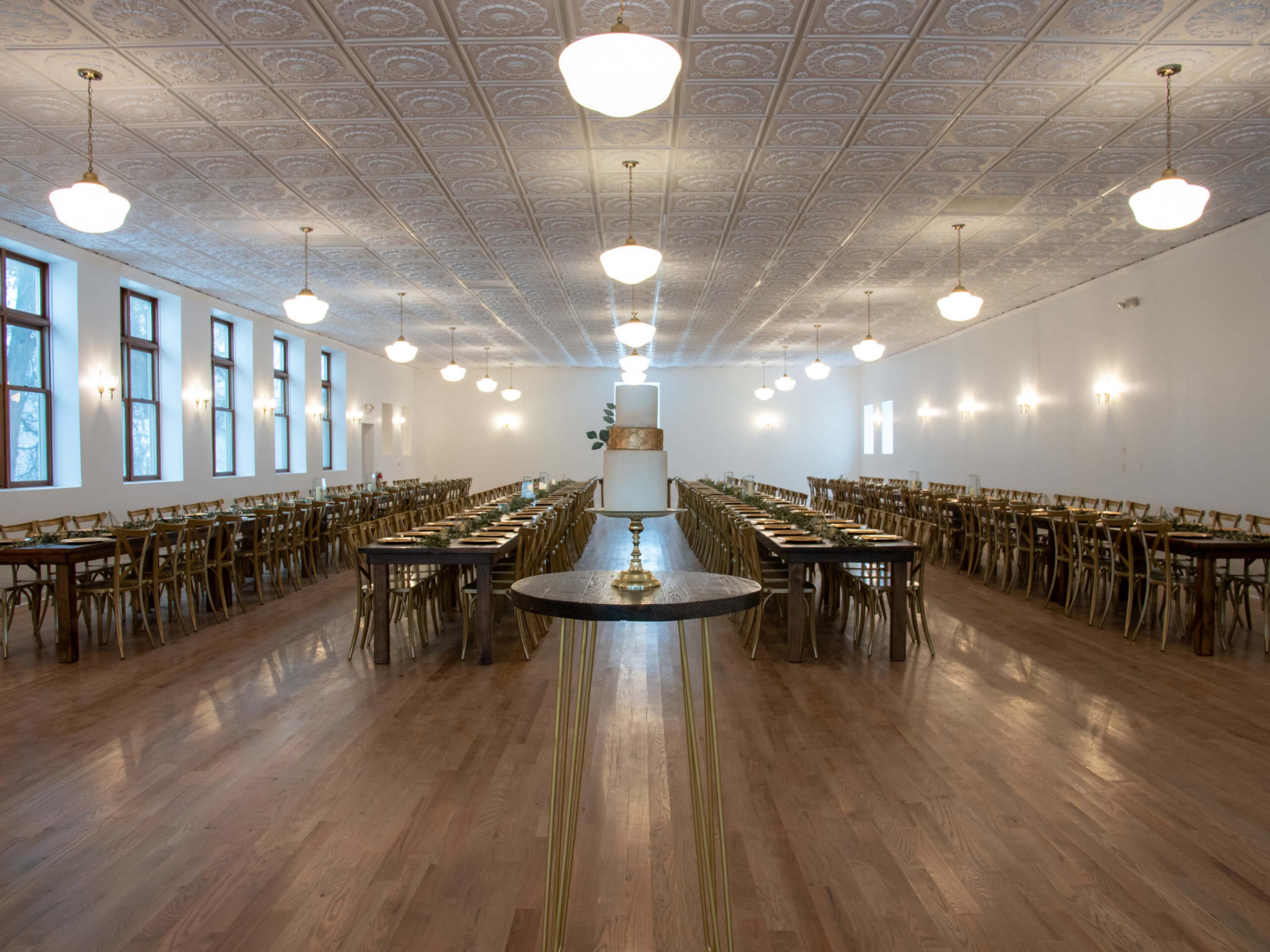 A large, elegantly arranged dining hall features multiple rows of tables and golden chairs, illuminated by overhead lights and large windows.
