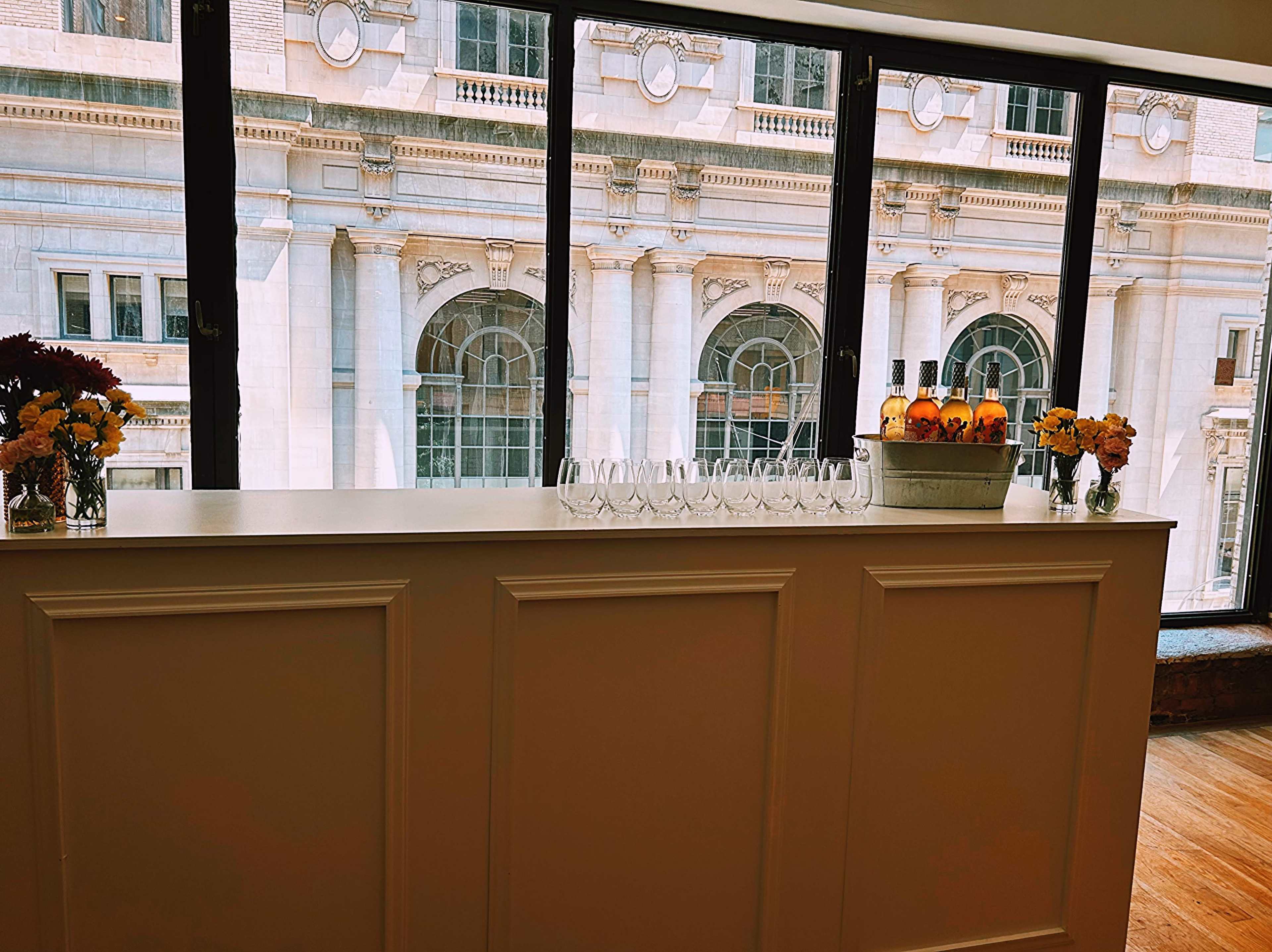 The image shows a modern indoor bar area with a row of wine glasses and a vase of flowers, overlooking a historic building through large windows.