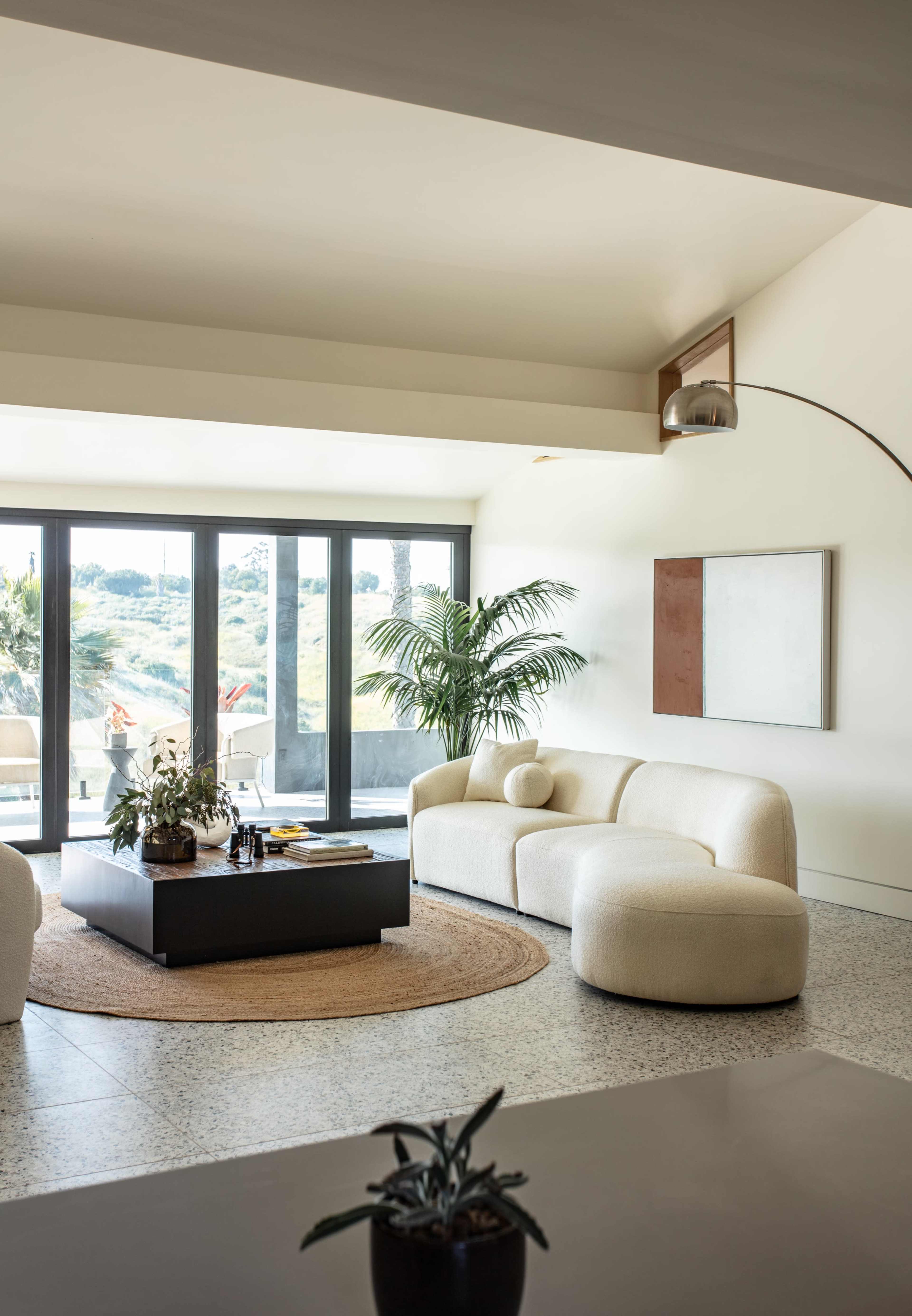 A spacious living room featuring a curved white sofa, a large black coffee table, and a potted plant, with wide glass doors providing a view of the outdoors.