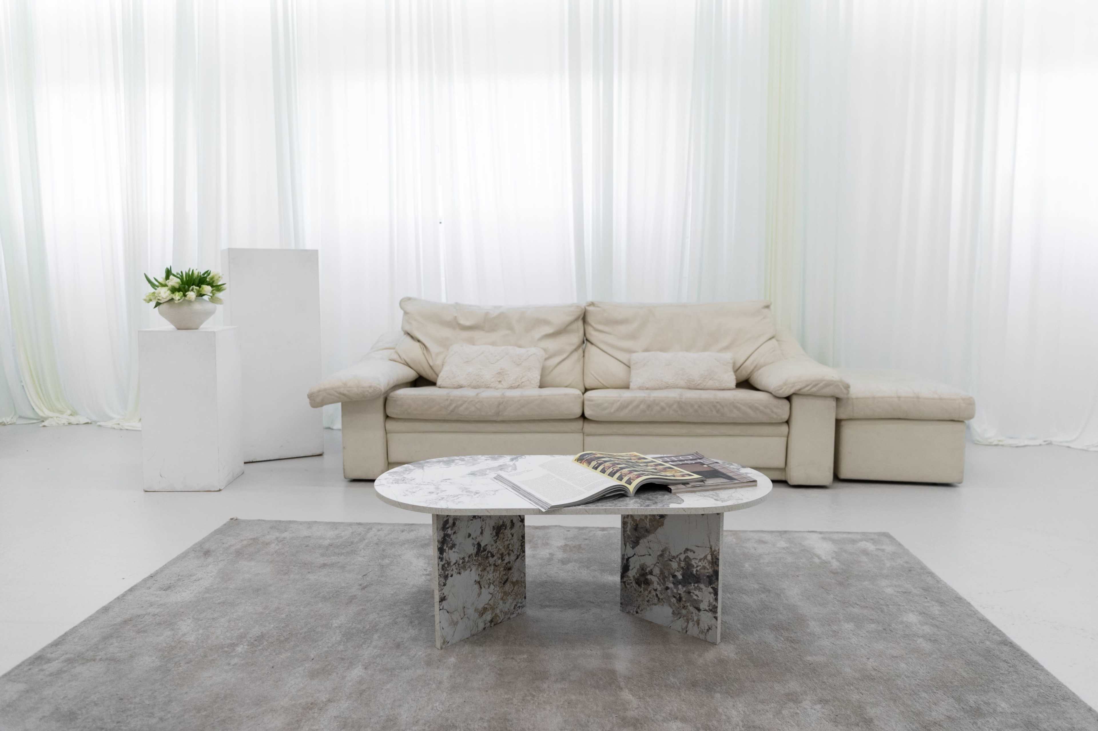 A beige sofa is positioned in a minimalistic room with white drapes, accompanied by a circular table displaying a magazine and decorated with a subtle rug underneath.