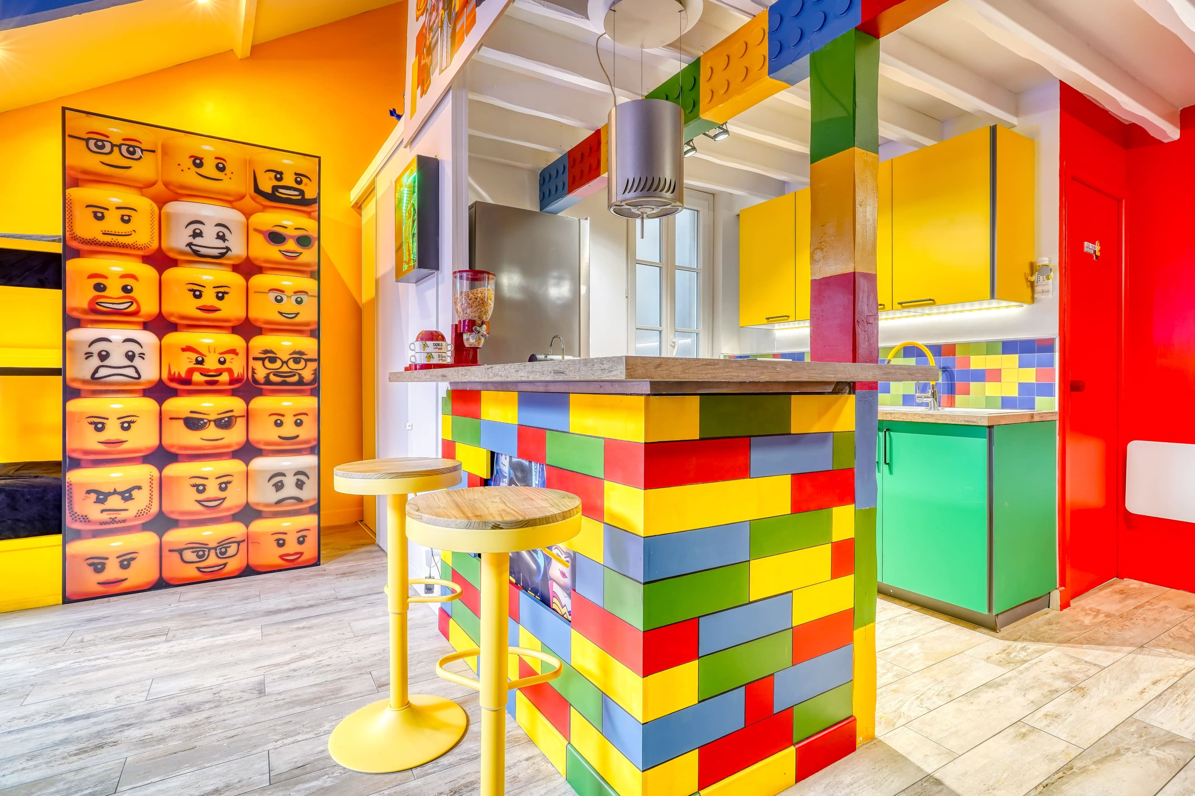 The image shows a brightly colored kitchen with walls and furniture featuring a Lego brick design, including a bar with yellow stools and a vibrant green cabinet.