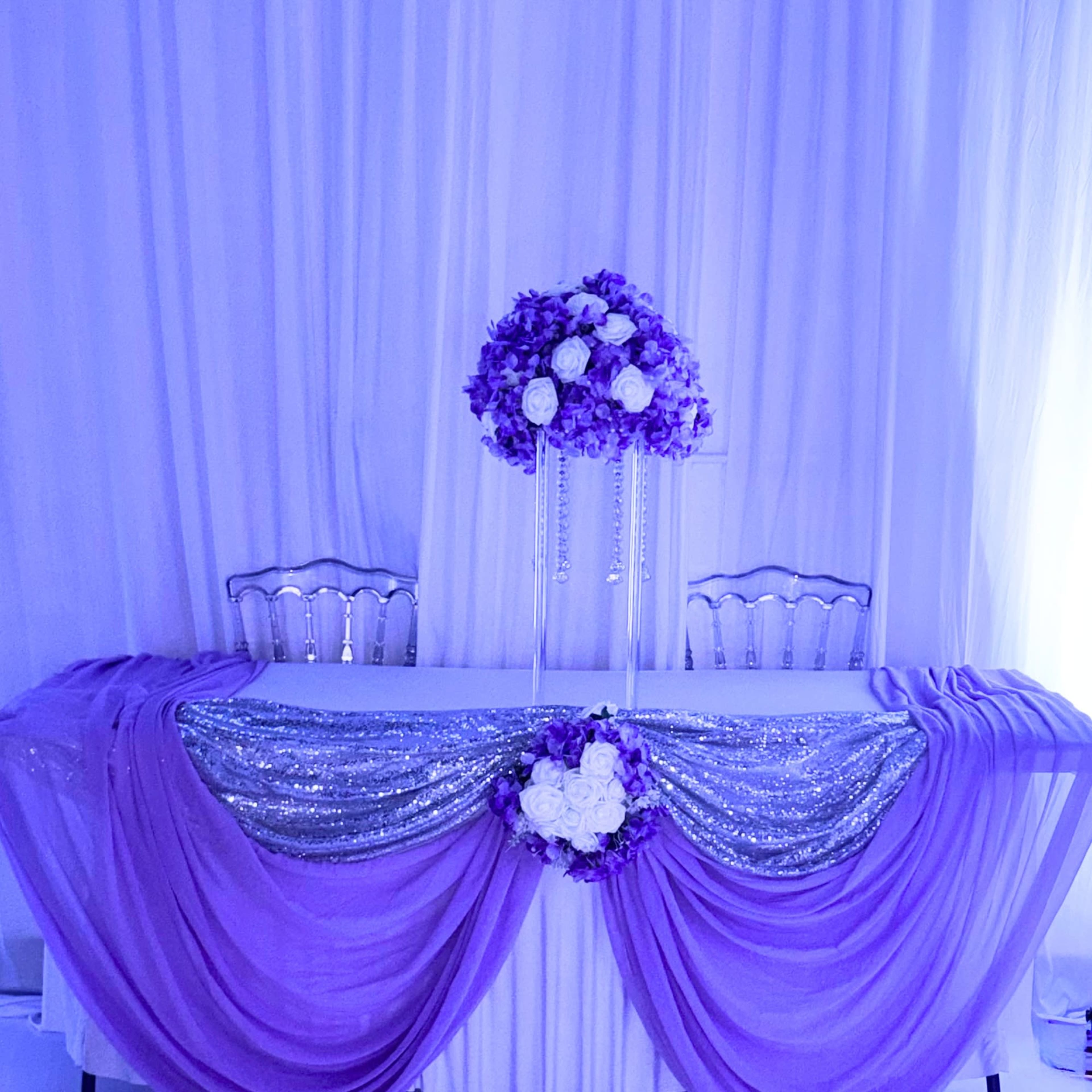 The image shows a decorated table draped in purple fabric, embellished with silver sequins and adorned with floral arrangements.