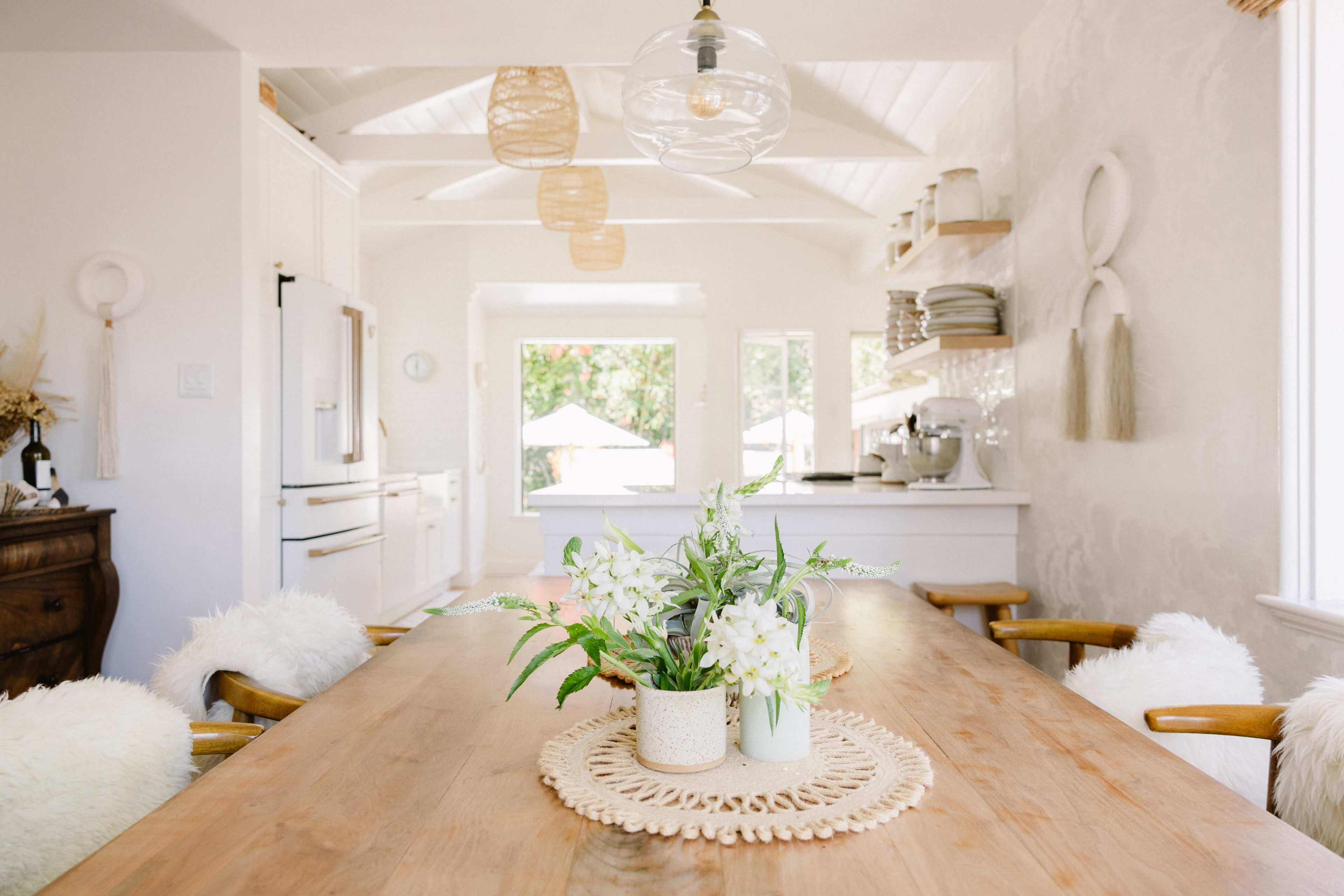 The image shows a bright, modern kitchen and dining area featuring a wooden table with a decorative centerpiece of flowers, surrounded by light-colored chairs.