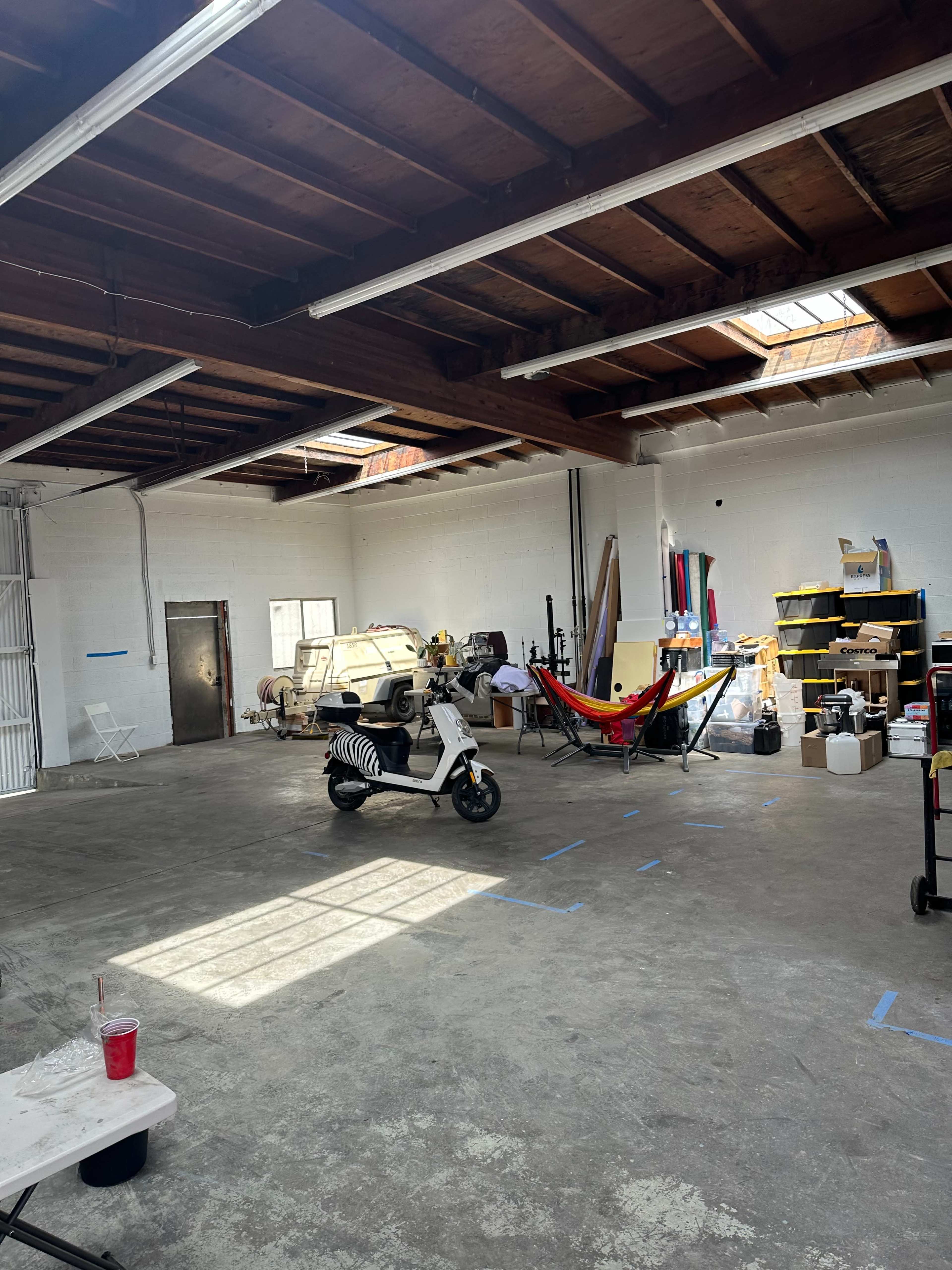 The image shows a spacious garage or workshop area containing various items, including a scooter, a hammock, and numerous boxes and tools scattered around.