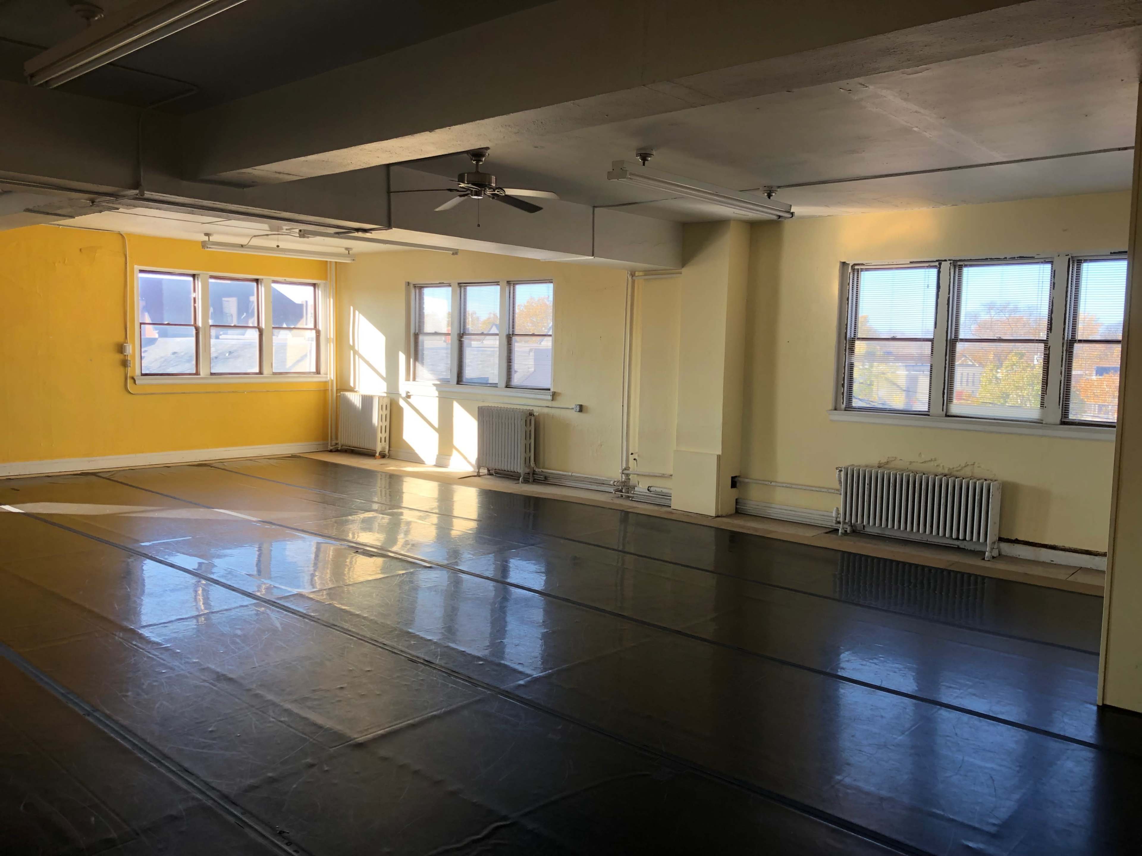 An empty room with a glossy black floor and large windows letting in natural light, painted yellow walls, and heating units along the walls.