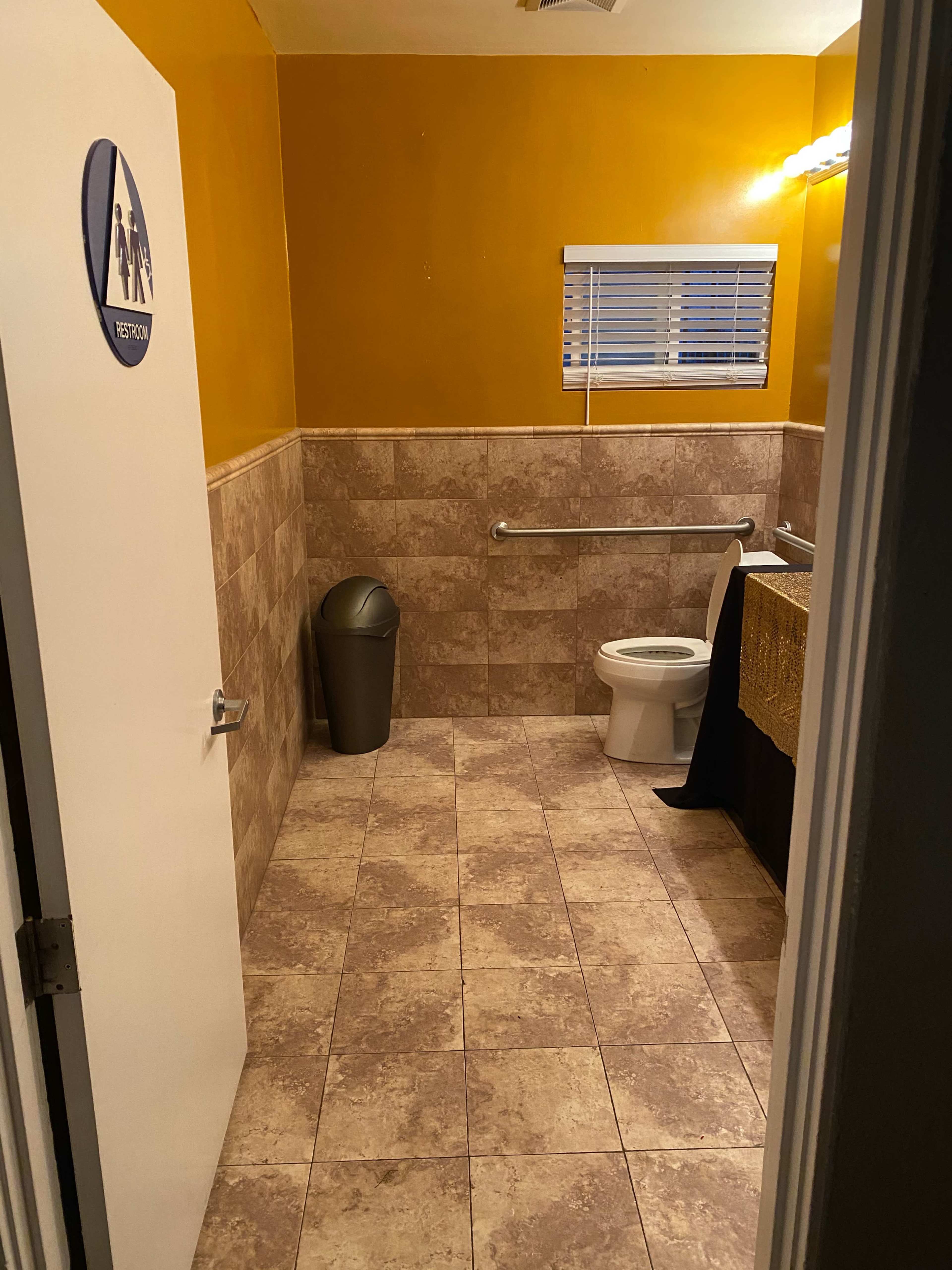 A small bathroom features beige tiled flooring, a toilet, a sink with a brown countertop, a trash can, and a door marked with a unisex restroom sign.