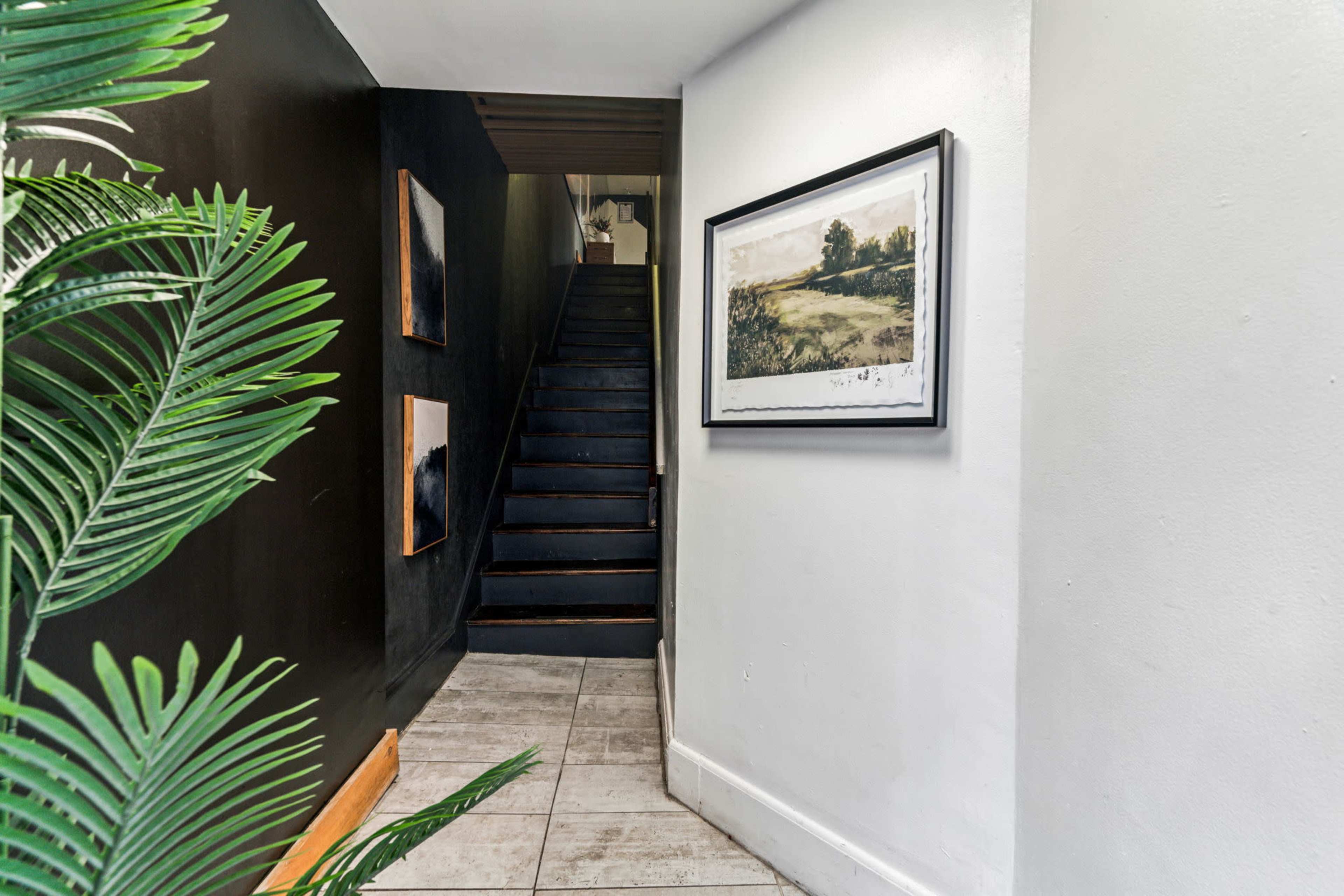 The image shows a hallway leading to a staircase, with a large framed painting on the wall and two smaller artworks beside it, along with a decorative plant in the foreground.