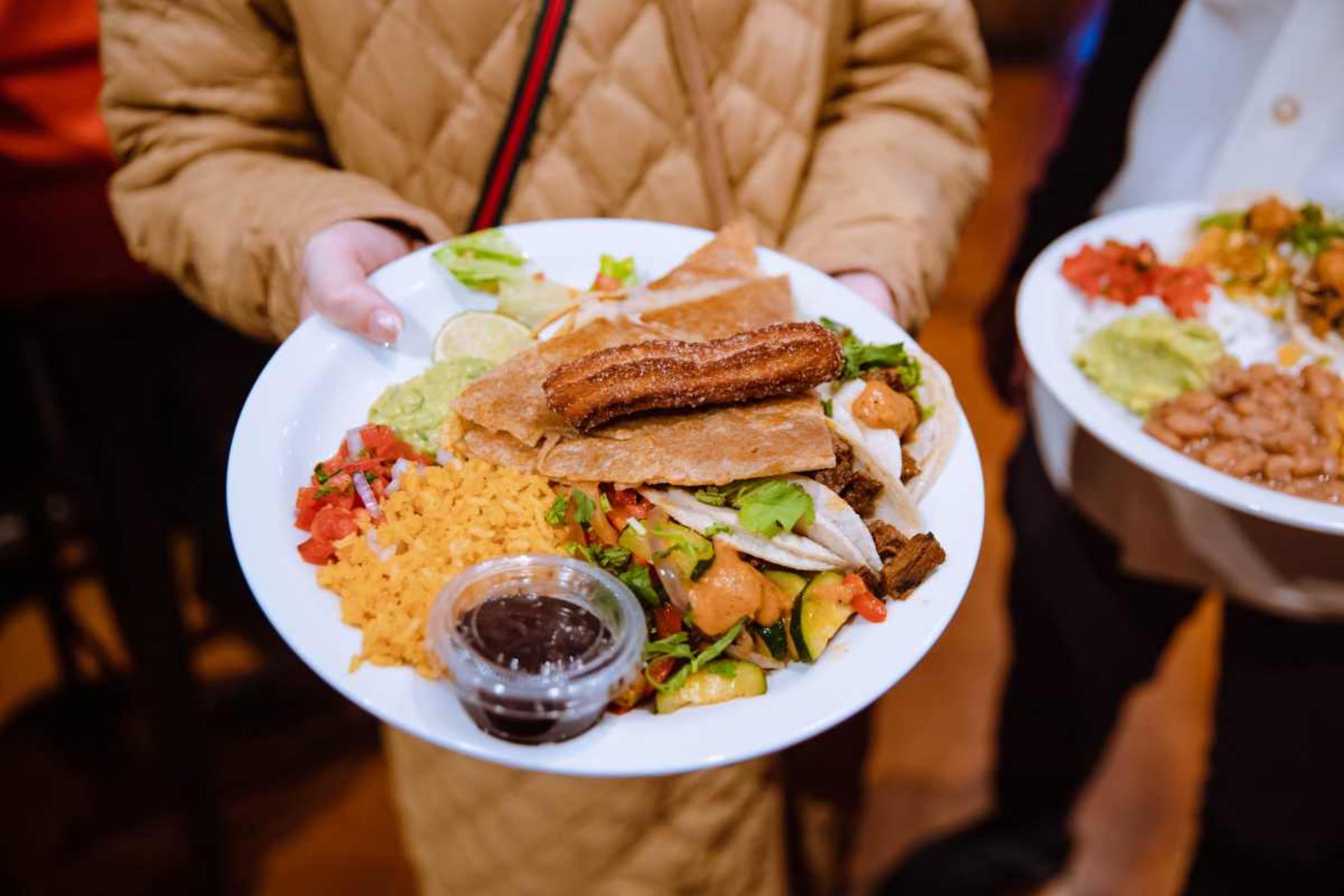 A person holds a plate of food featuring a variety of items including beans, rice, salad, tortilla chips, guacamole, and a fried item.