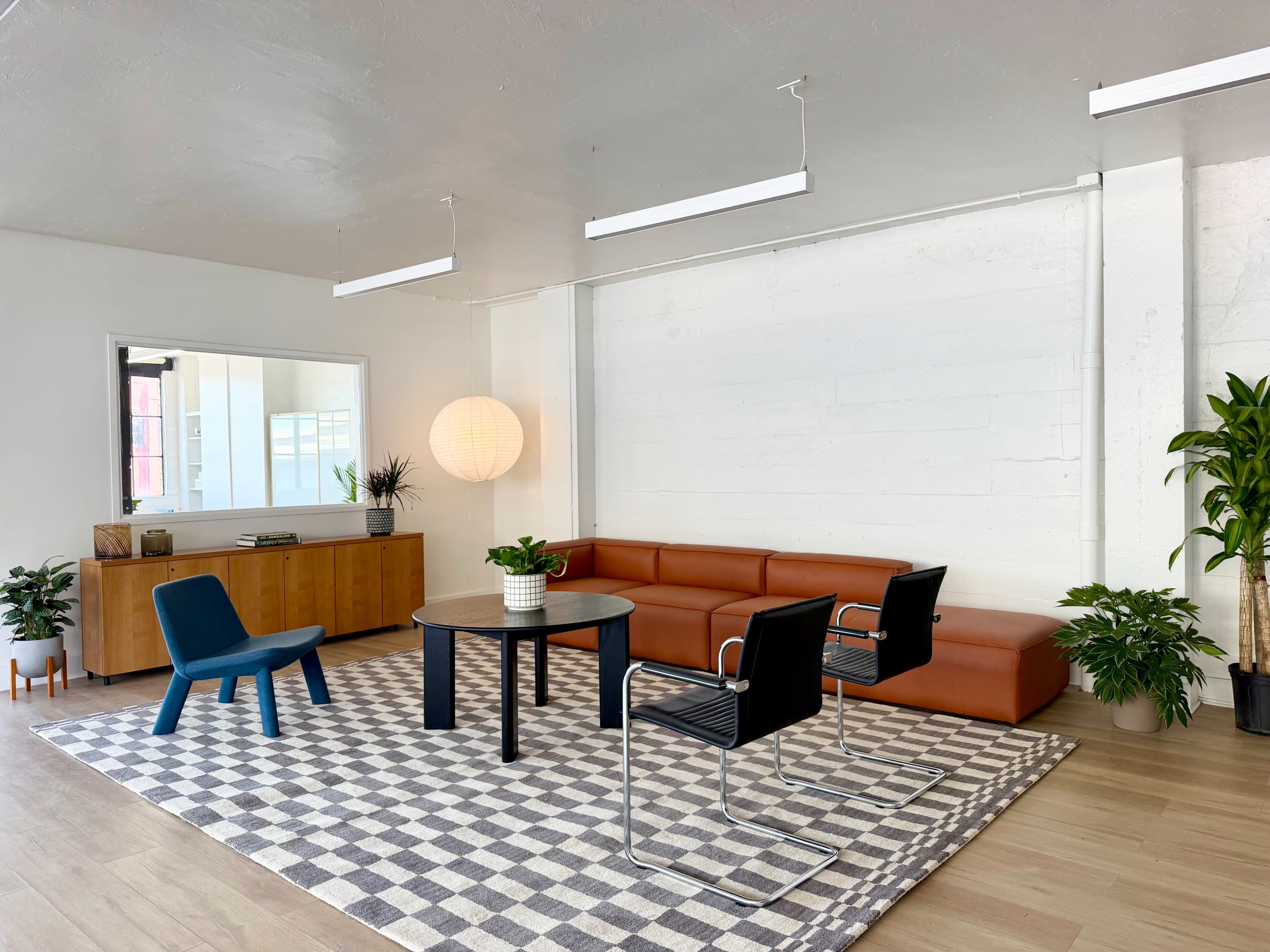 The image shows a modern, minimalist living space with a brown leather sofa, a round table surrounded by black chairs, and a patterned rug on a wooden floor.