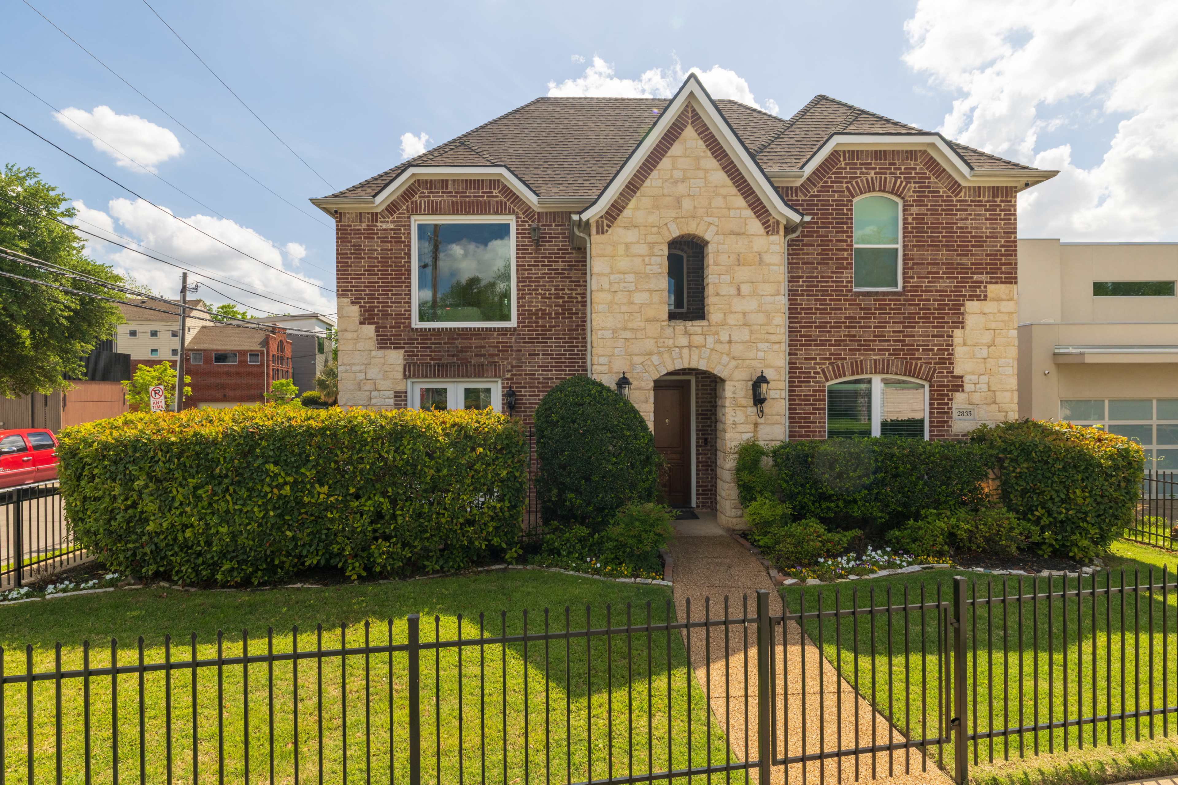 The image shows a two-story brick house with stone accents, surrounded by a green hedge and a black metal fence.