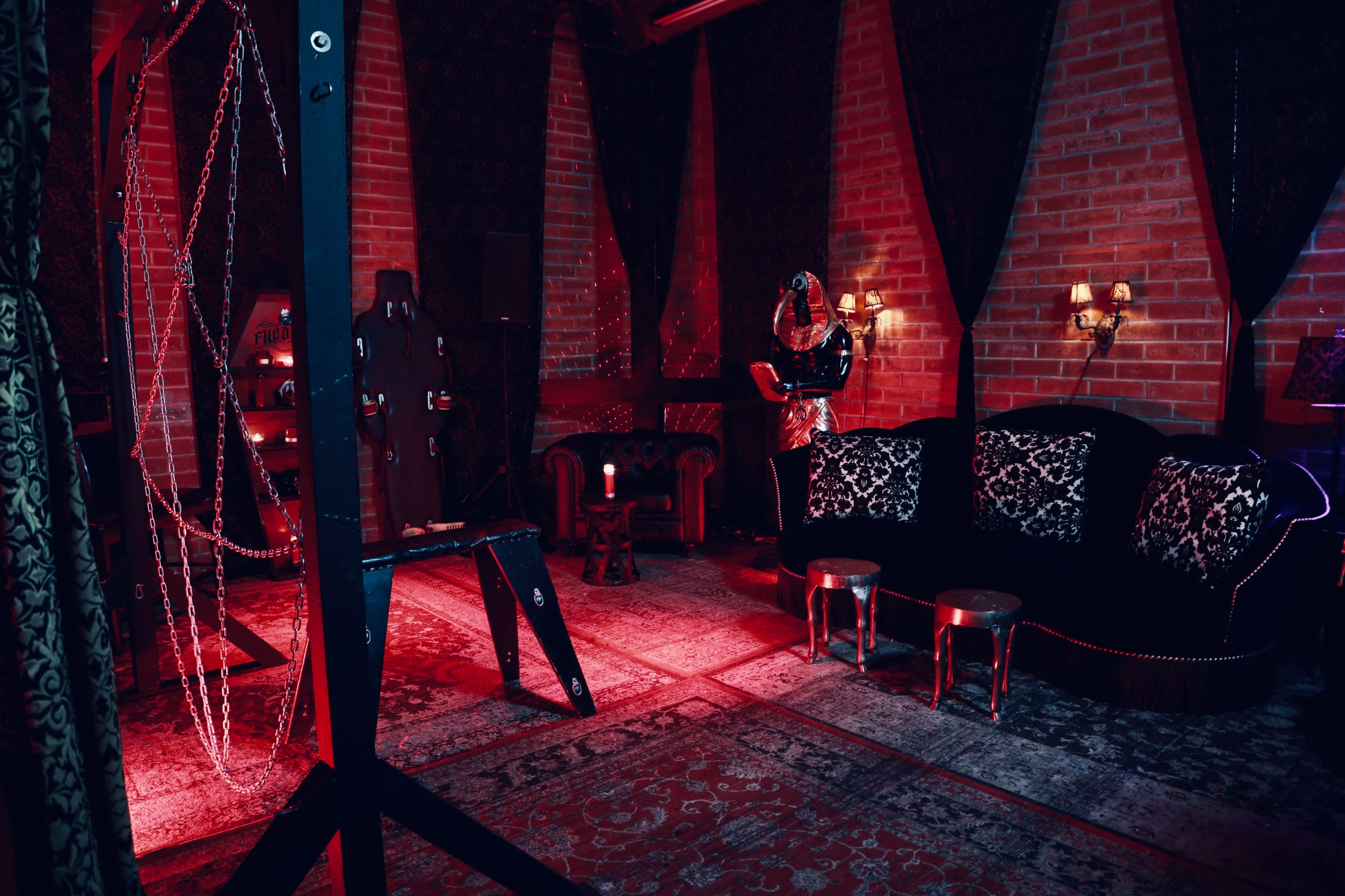 The room features dark walls adorned with drapes, a vintage sofa, small tables, and various BDSM equipment arranged around a patterned rug.