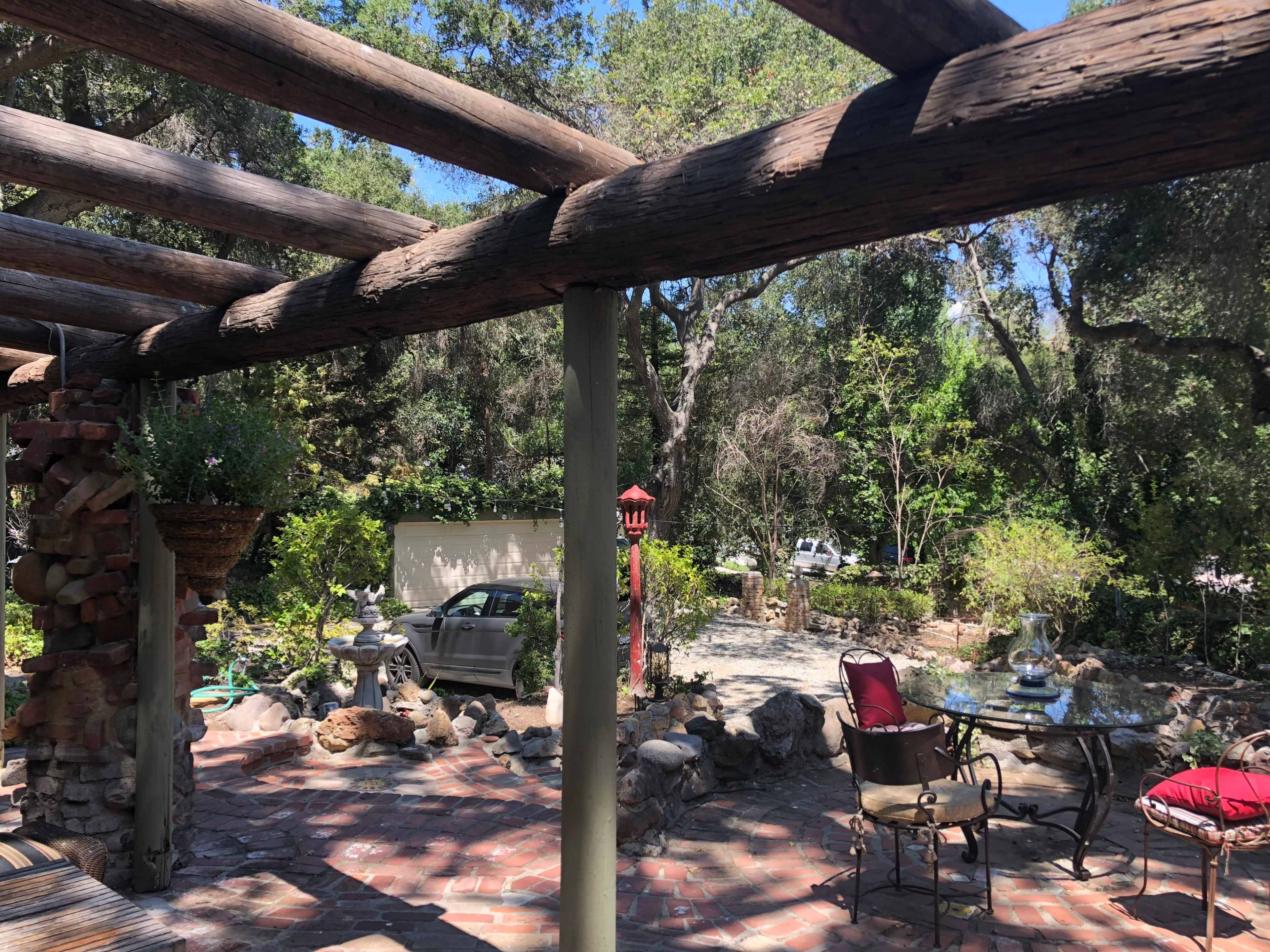 A stone patio is framed by wooden beams, overlooking a garden area with various plants and a parked car in the background.