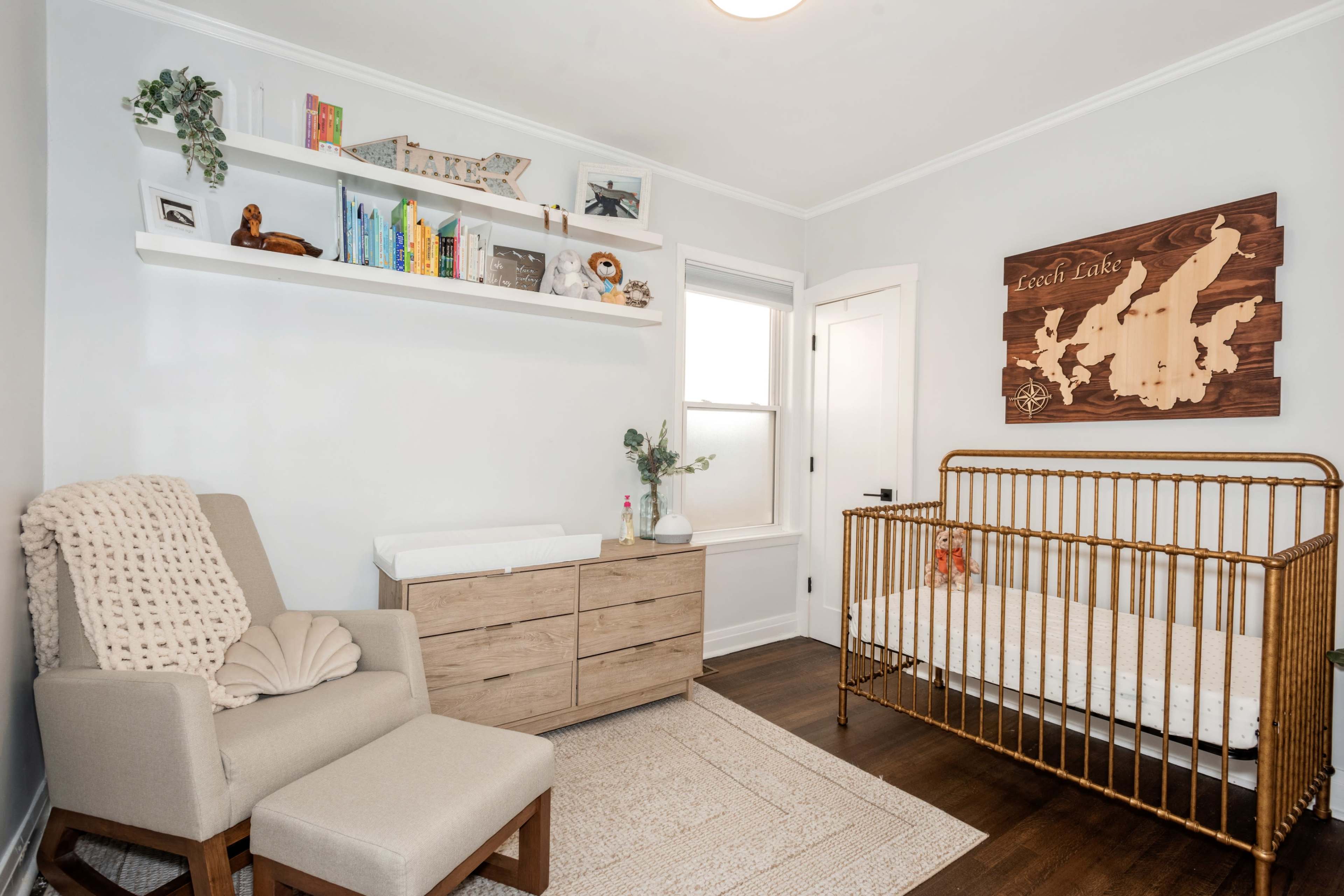 A nursery features a wooden crib, a stylish rocking chair, a changing table, and shelves with children's books and decorative items.