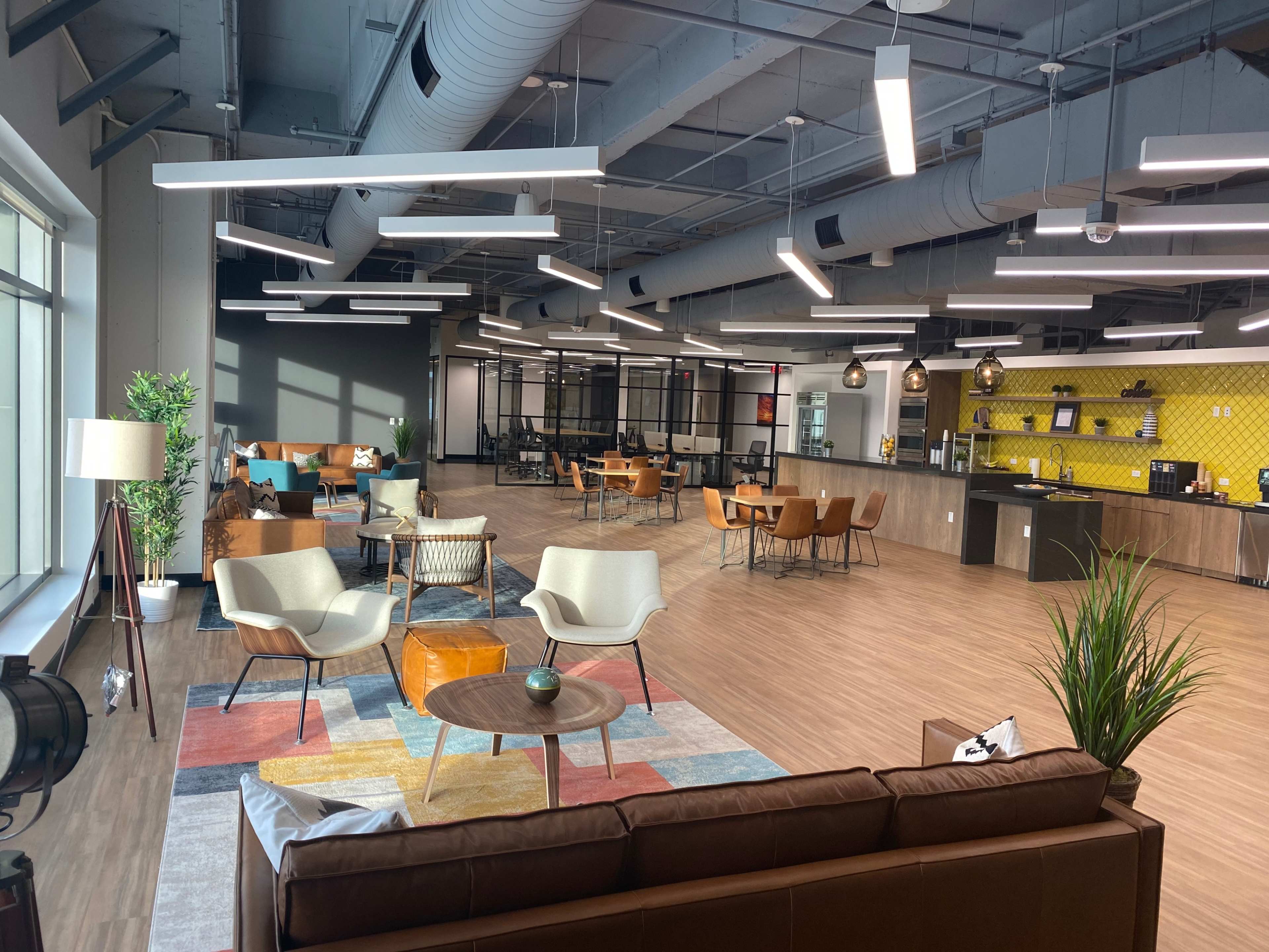 A modern open office space with seating areas, a kitchenette, and large windows providing natural light.