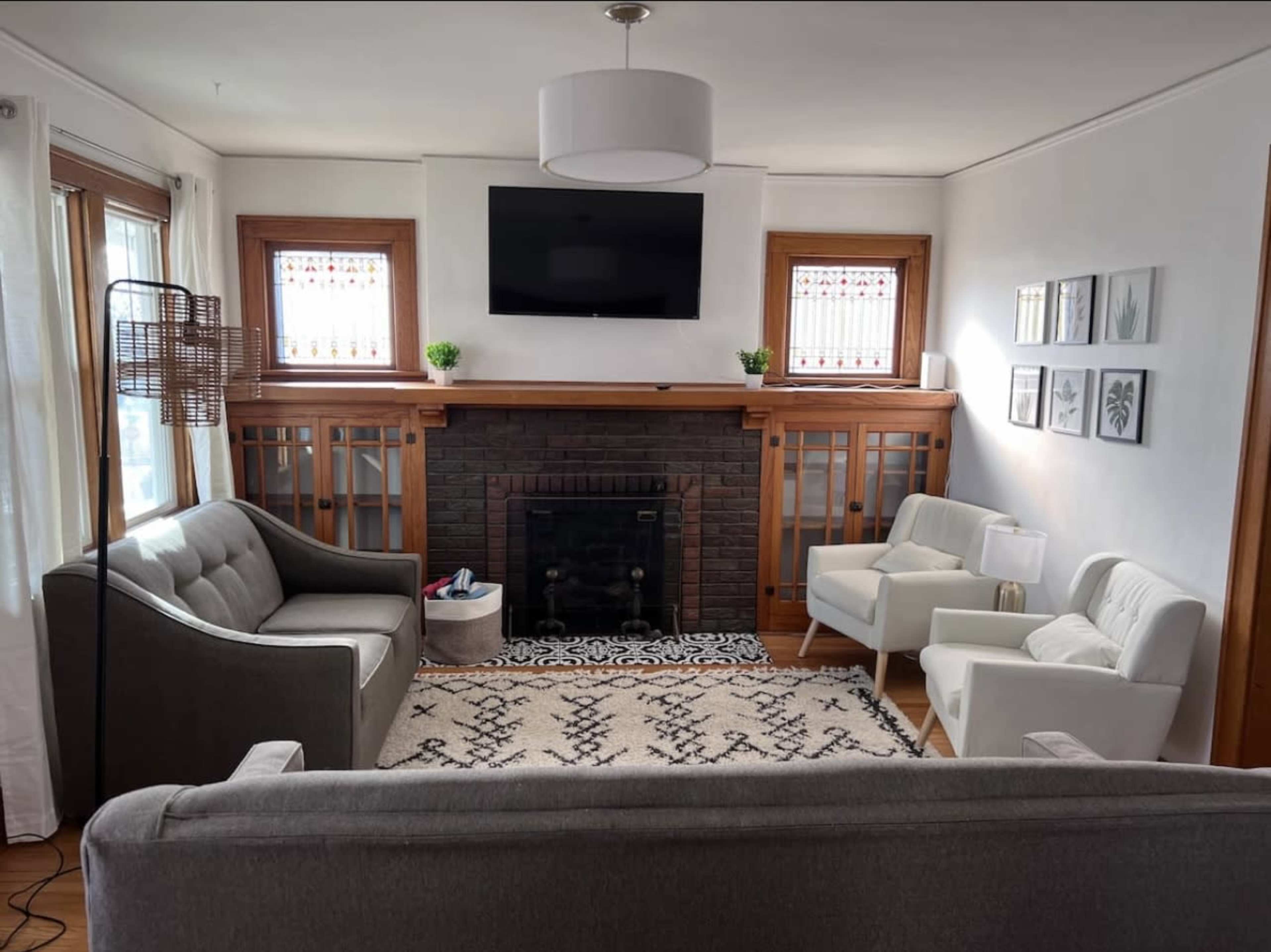 A living room features a gray sofa, two white armchairs, a black fireplace with a television above it, and decorative plants on the shelves.