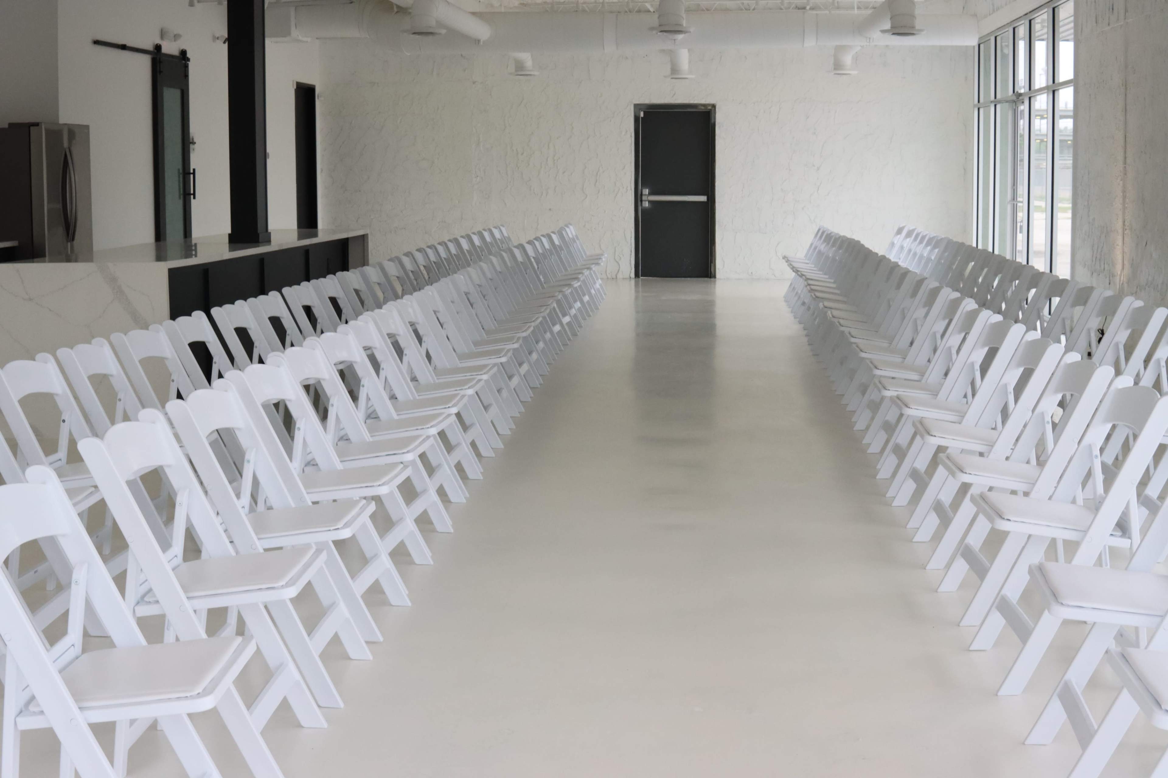A long, empty room with two rows of white folding chairs facing each other, creating a pathway in the center.