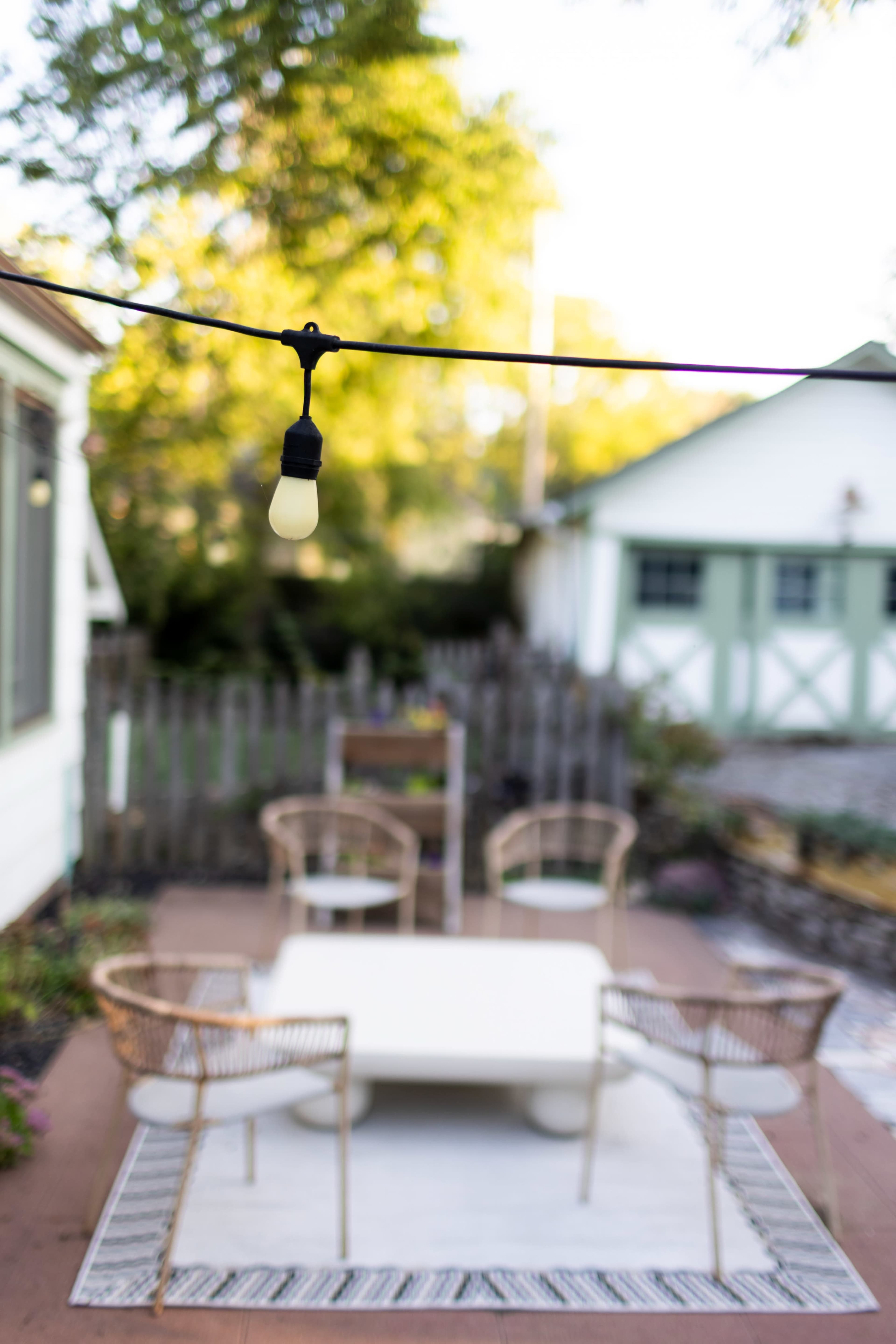 A string light hangs above a patio area with four chairs surrounding a white table and a backdrop of a green shed.
