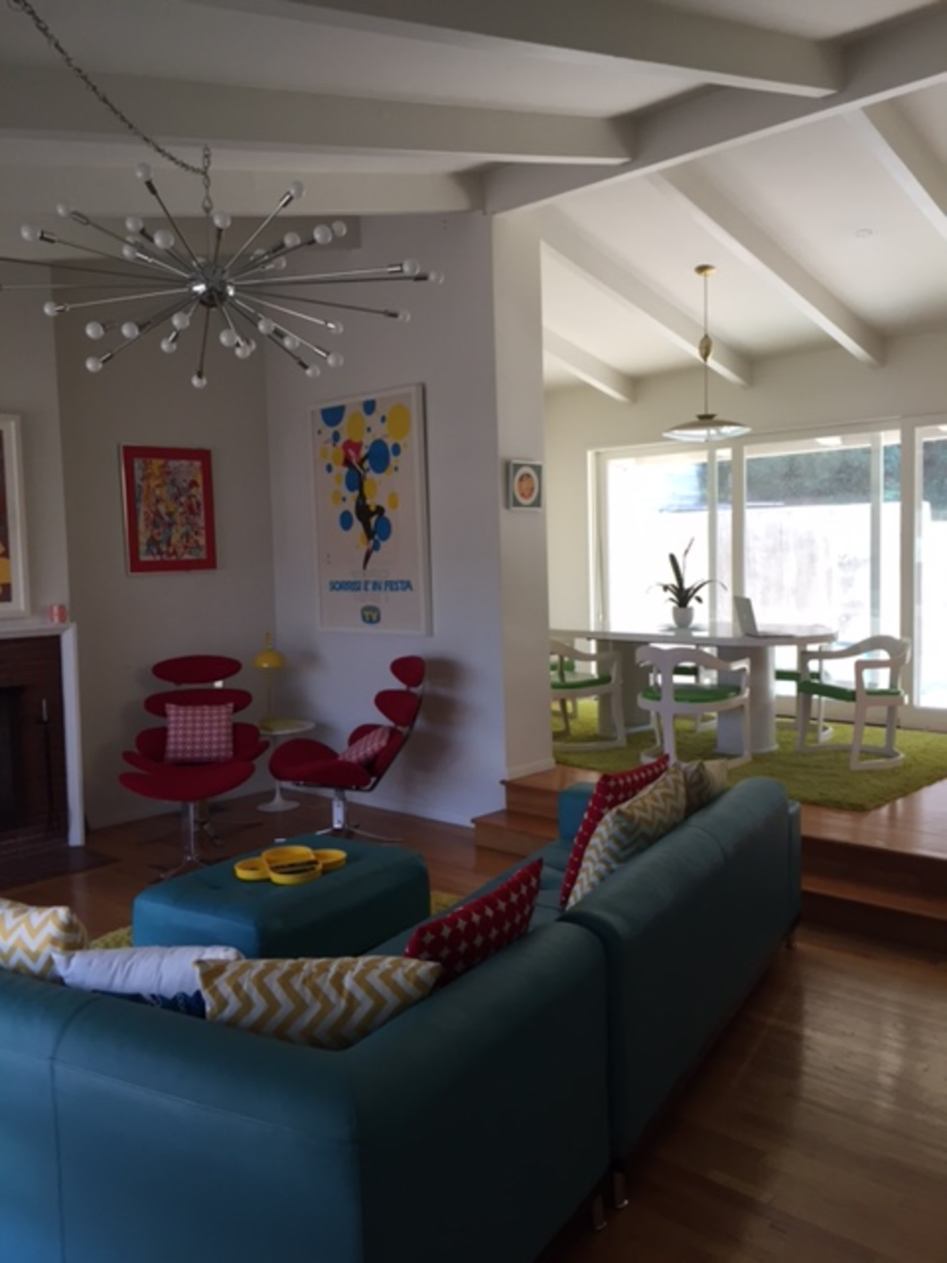 The image shows a modern living room with a turquoise sofa, red chairs, and a dining area featuring a table and chairs, accented by large windows and colorful artwork.