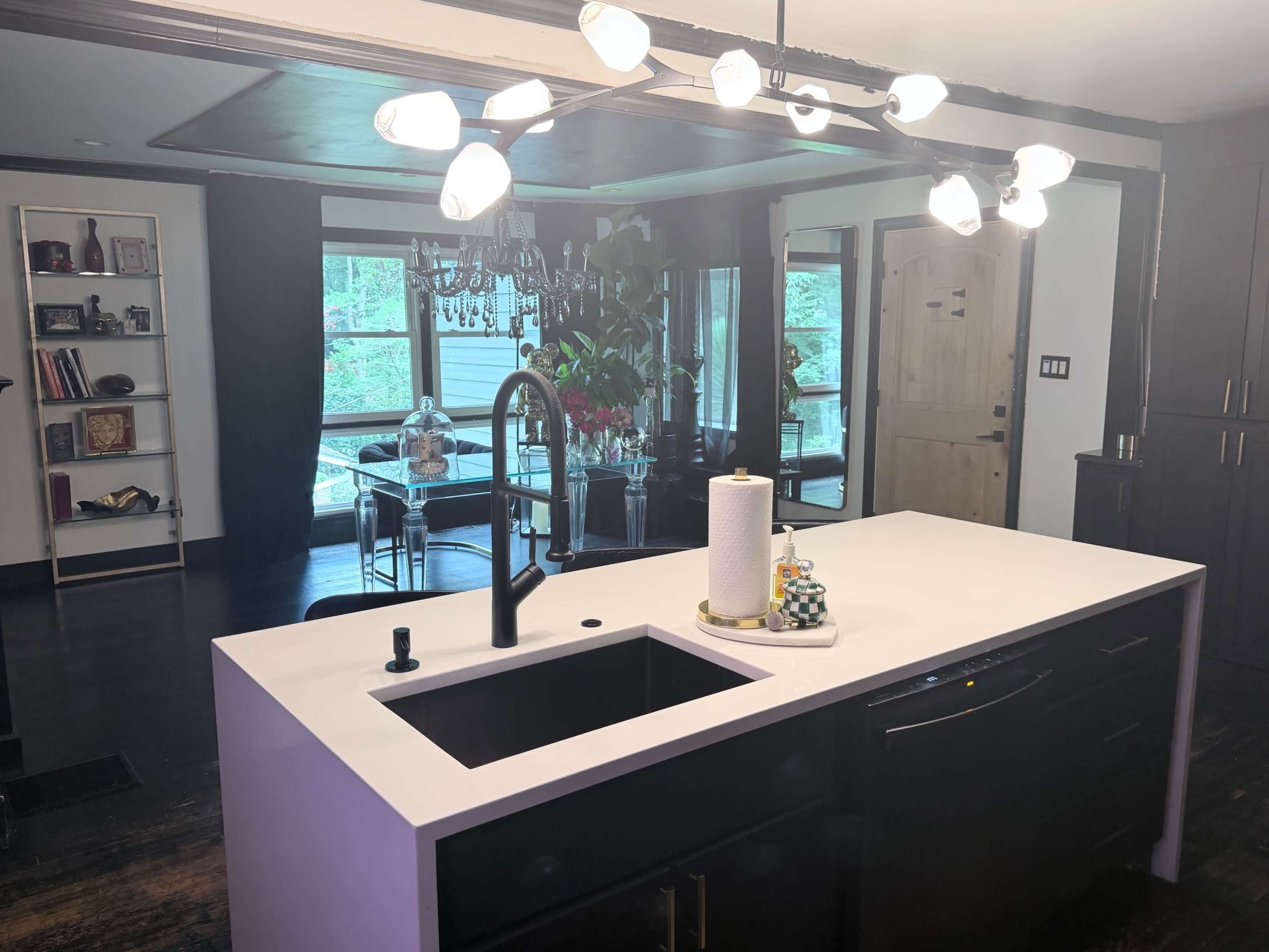 A modern kitchen features a black island with a sink and a stylish light fixture above, overlooking a dining area with a chandelier and large windows.