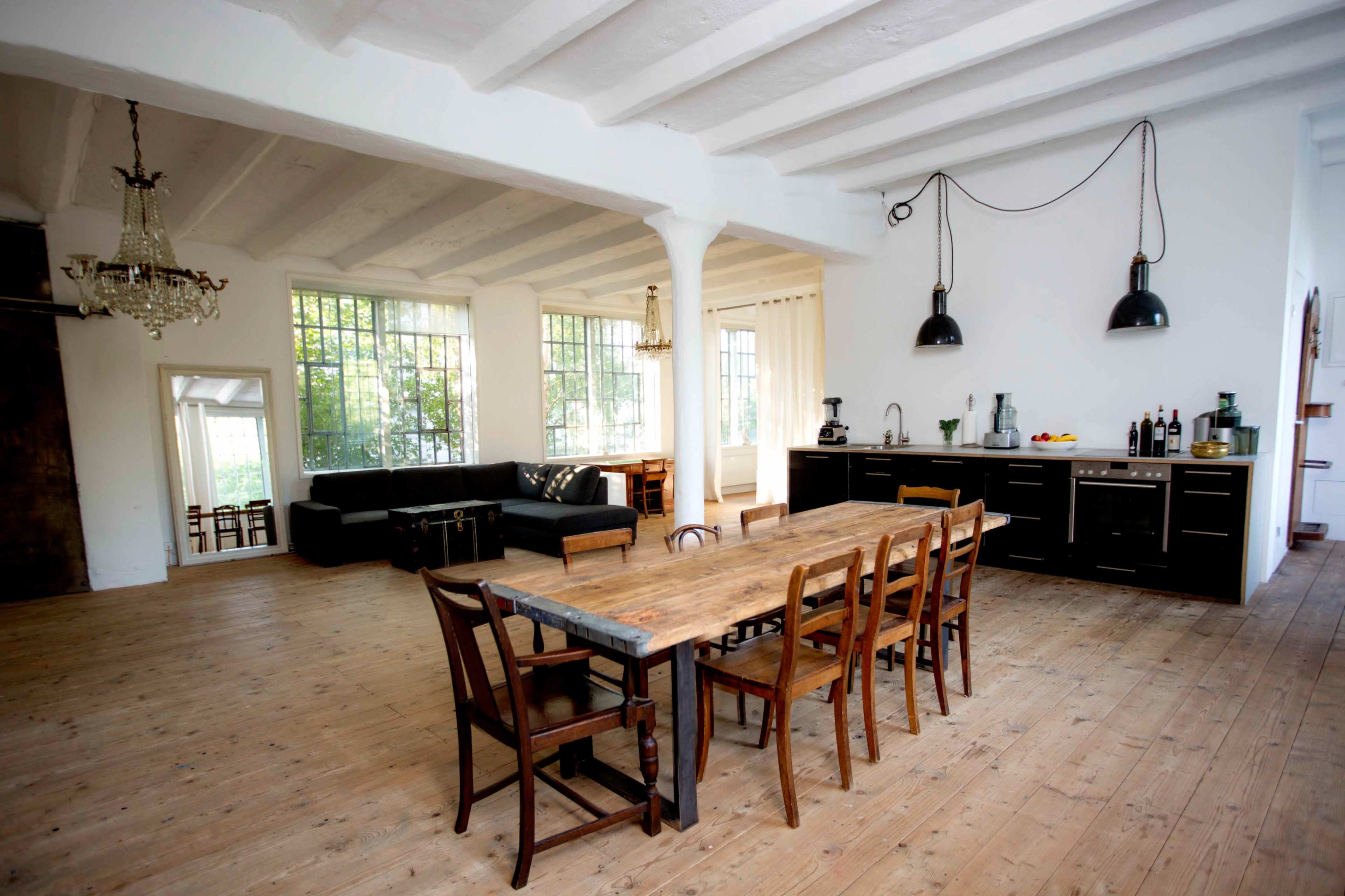 The image shows a spacious, well-lit open-concept kitchen and dining area featuring a wooden dining table with chairs, a black kitchen with modern appliances, and large windows letting in natural light.