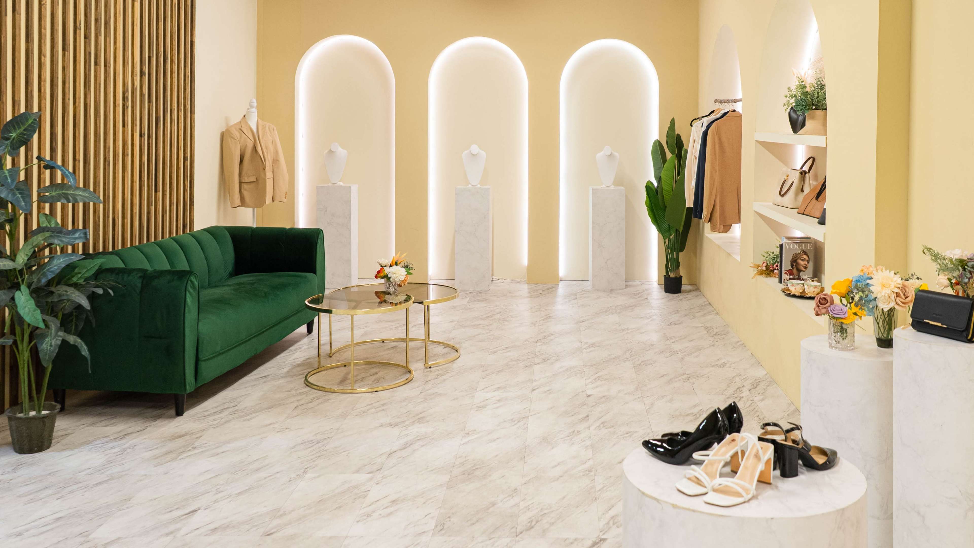 The image shows a stylish boutique interior with a green velvet sofa, marble display pedestals featuring clothing and accessories, and minimalist decor against a yellow wall.
