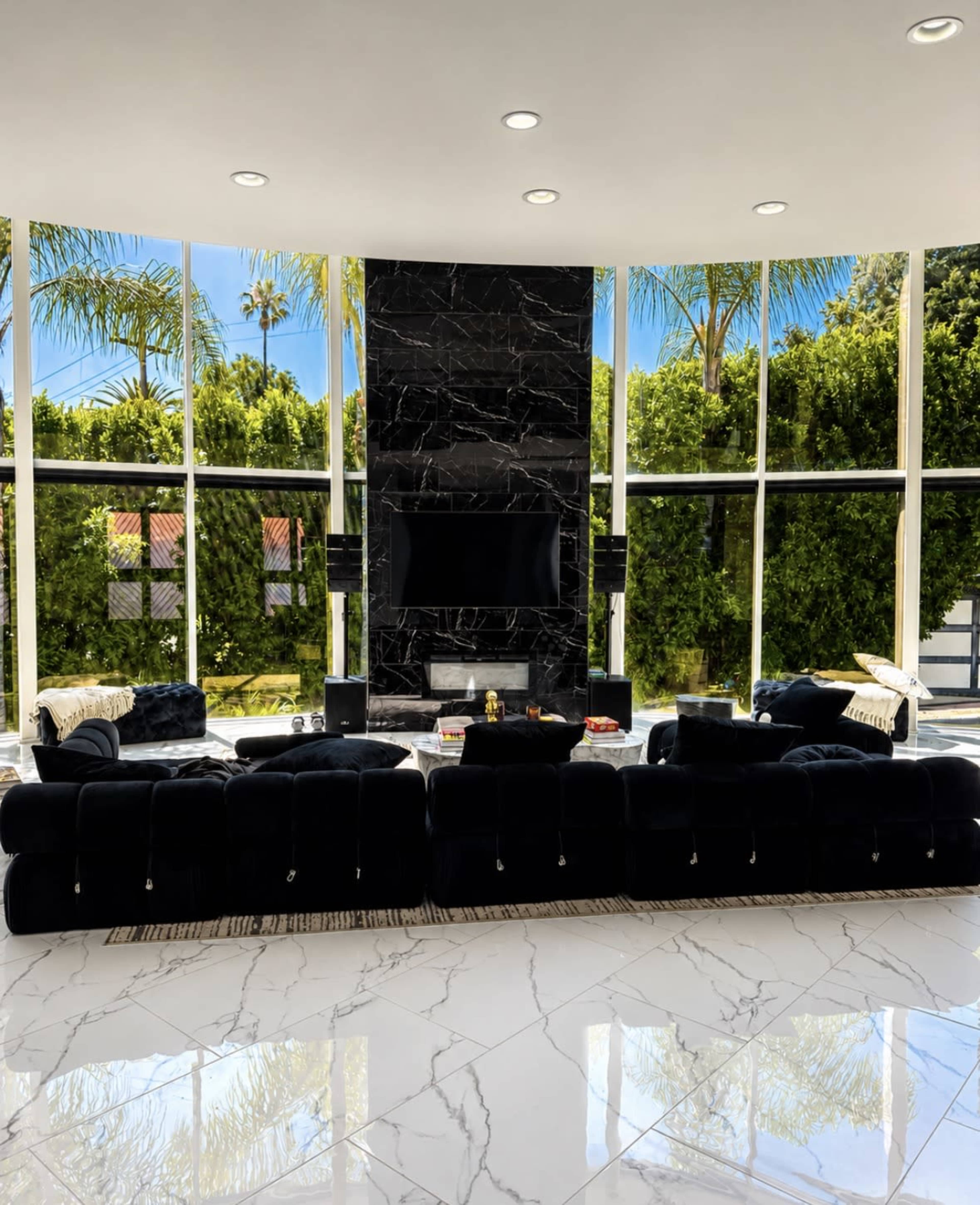 The image shows a modern living room with a large black sectional sofa, a sleek marble floor, and a wall-mounted television surrounded by large windows offering a view of lush greenery.