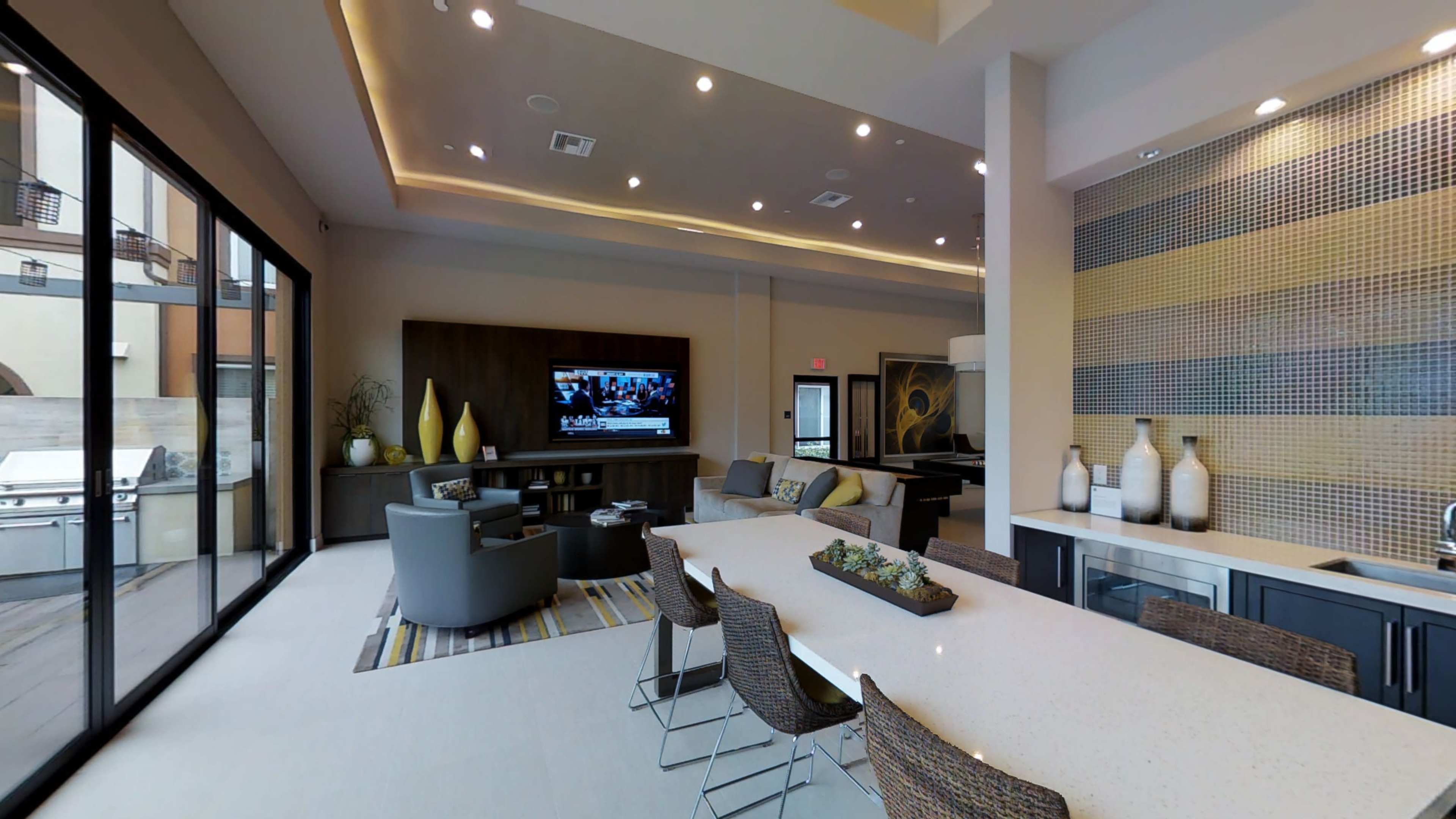 The image shows a modern lounge area with a large dining table, comfortable seating, a television on the wall, and large windows providing natural light.