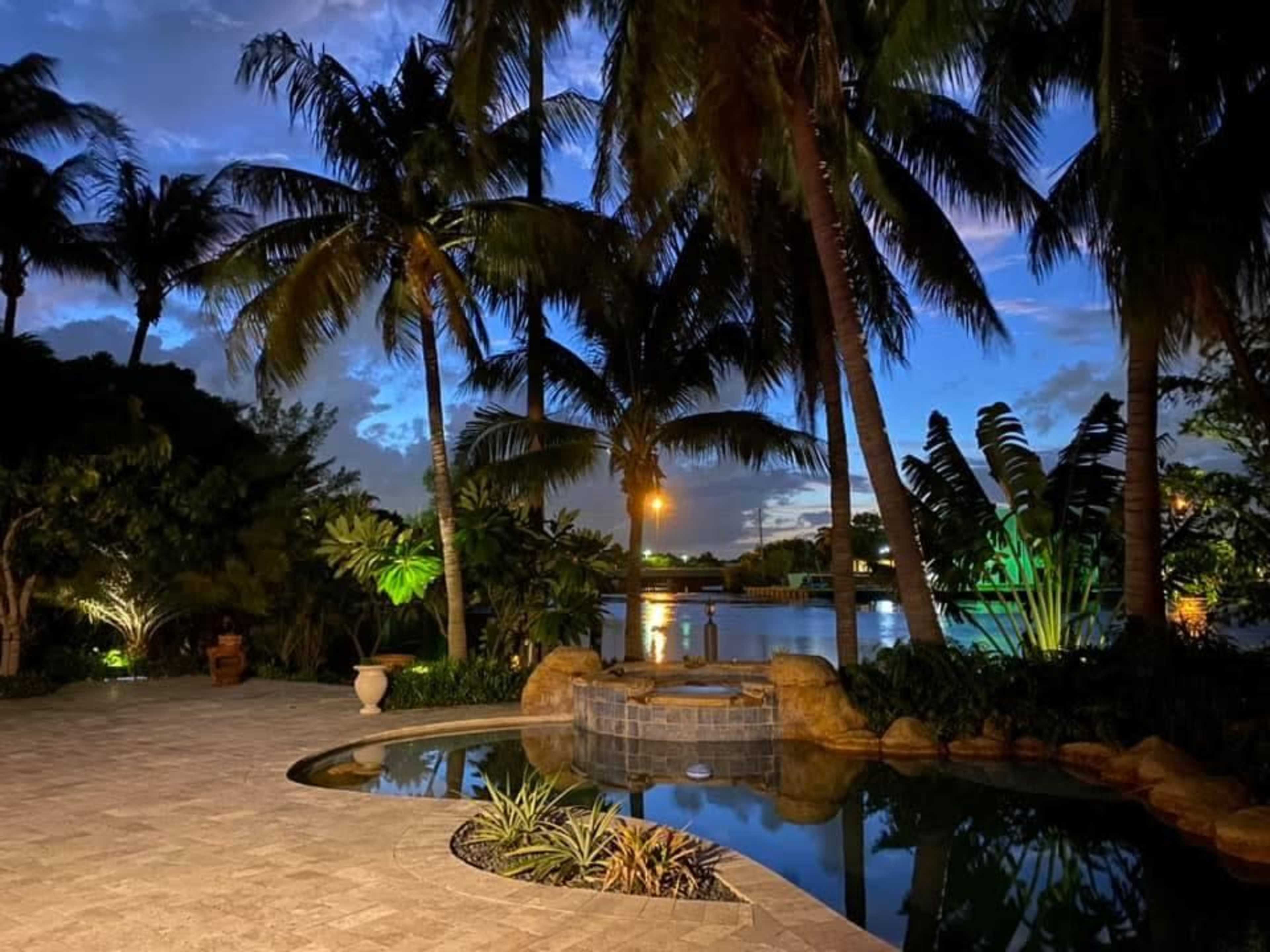 The scene features a tropical landscape with palm trees surrounding a pool and spa, near a body of water under a twilight sky.