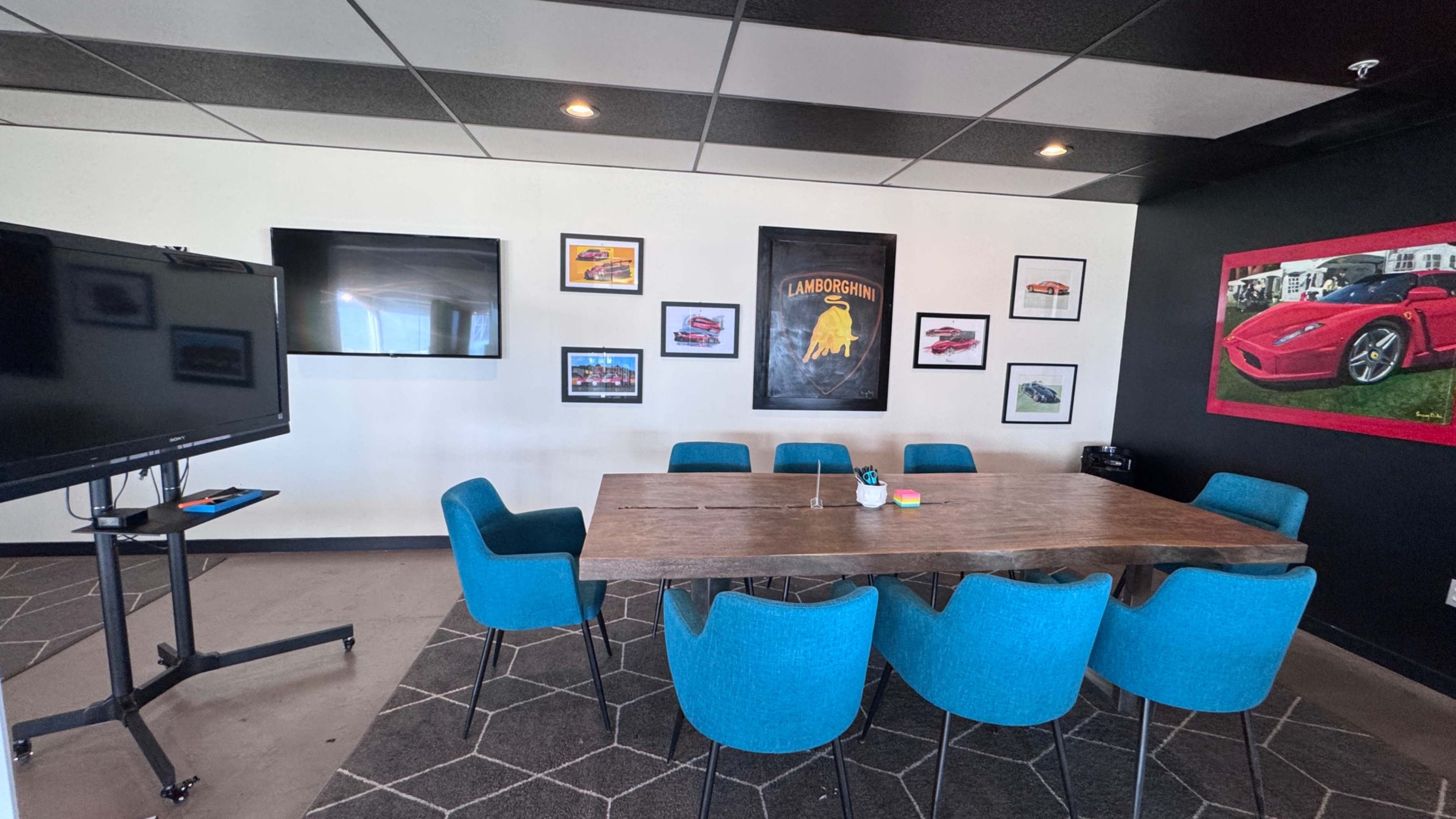 The image shows a modern conference room with a large wooden table surrounded by turquoise chairs, a television on a stand, and framed automotive art on the walls.