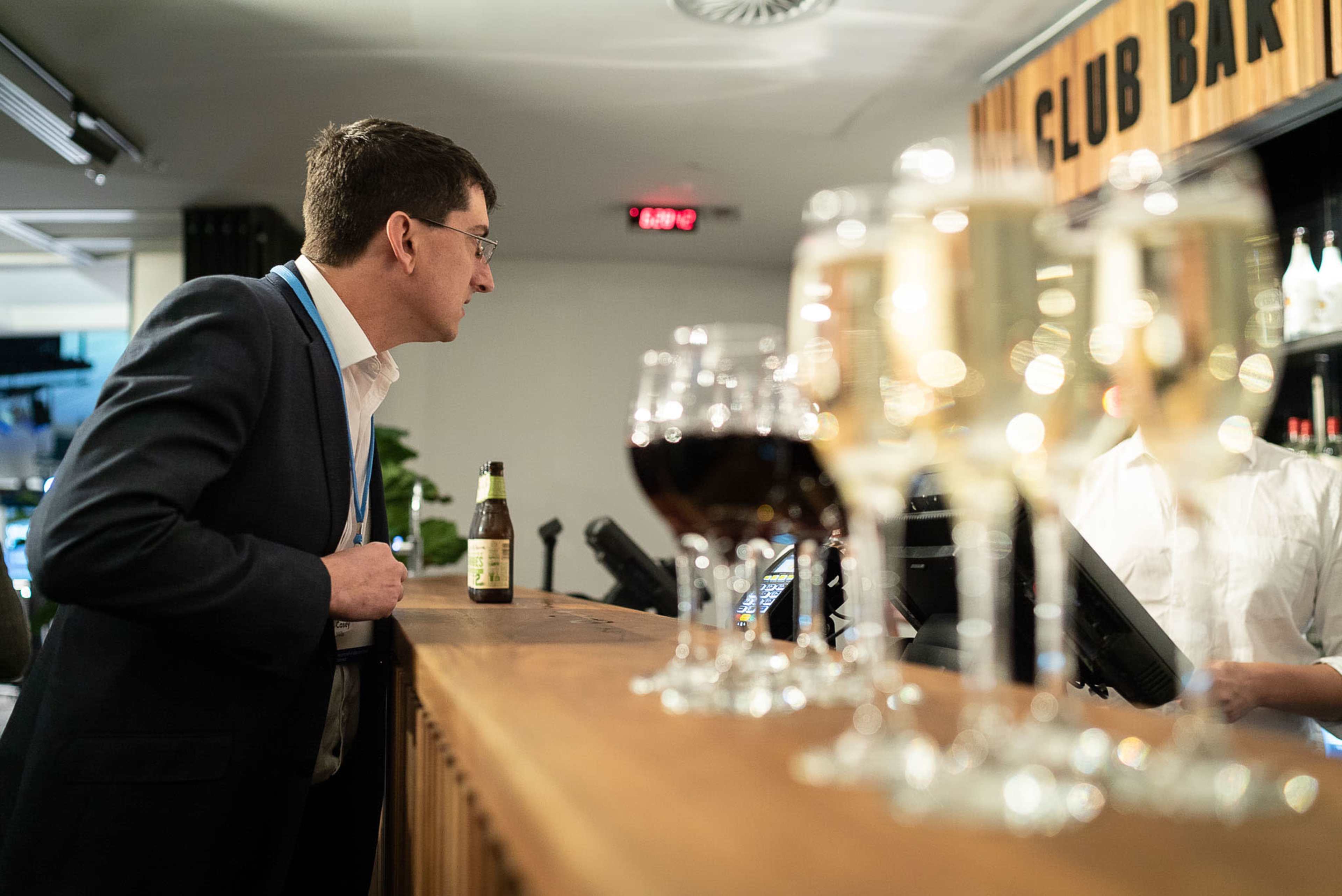 A man in a suit leans against a bar while observing the bartender prepare drinks, with various glasses of beverages lined up in the foreground.