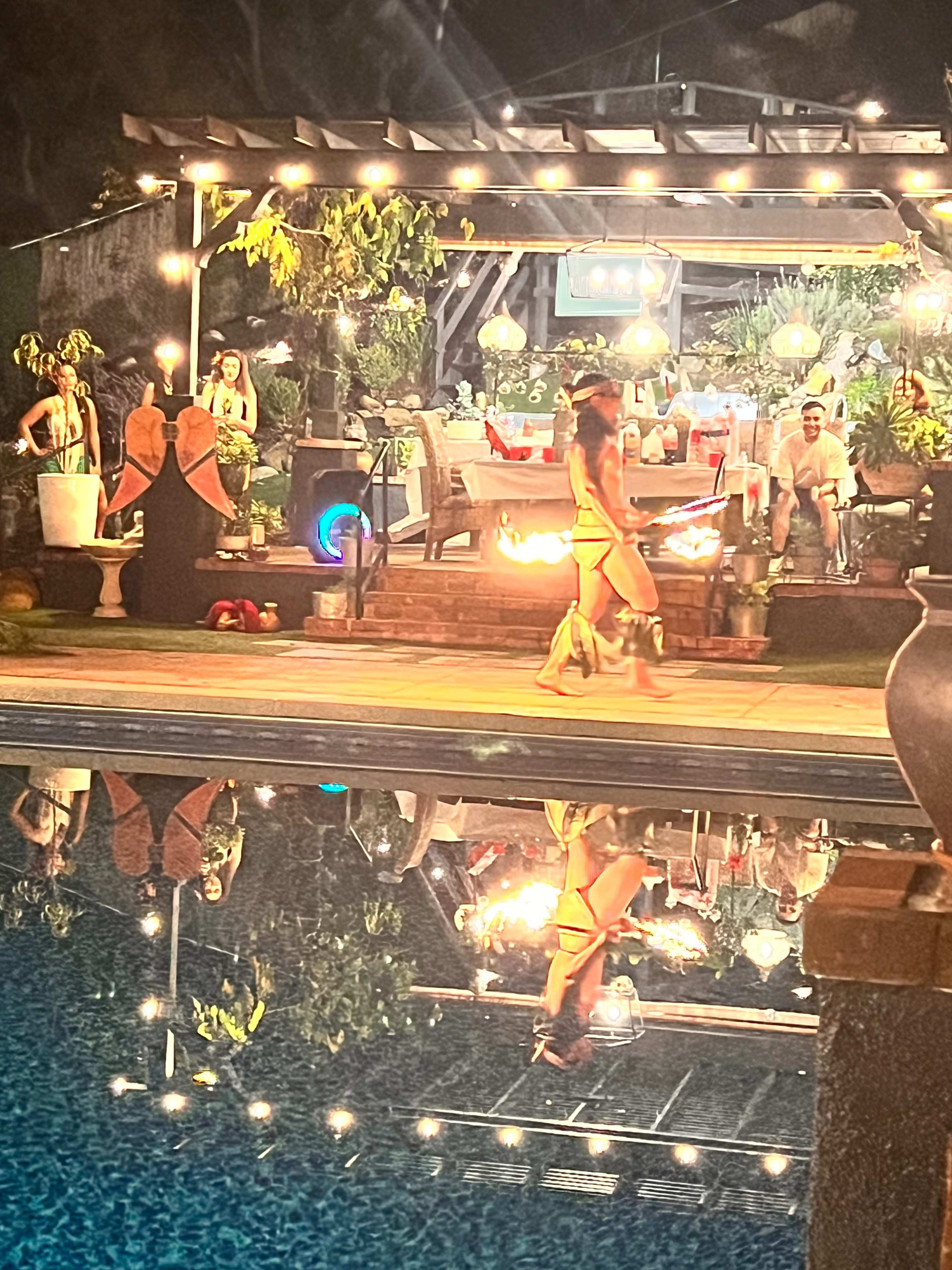 A woman twirls flaming poi beside a pool reflecting lights and a decorated outdoor space filled with people and greenery.