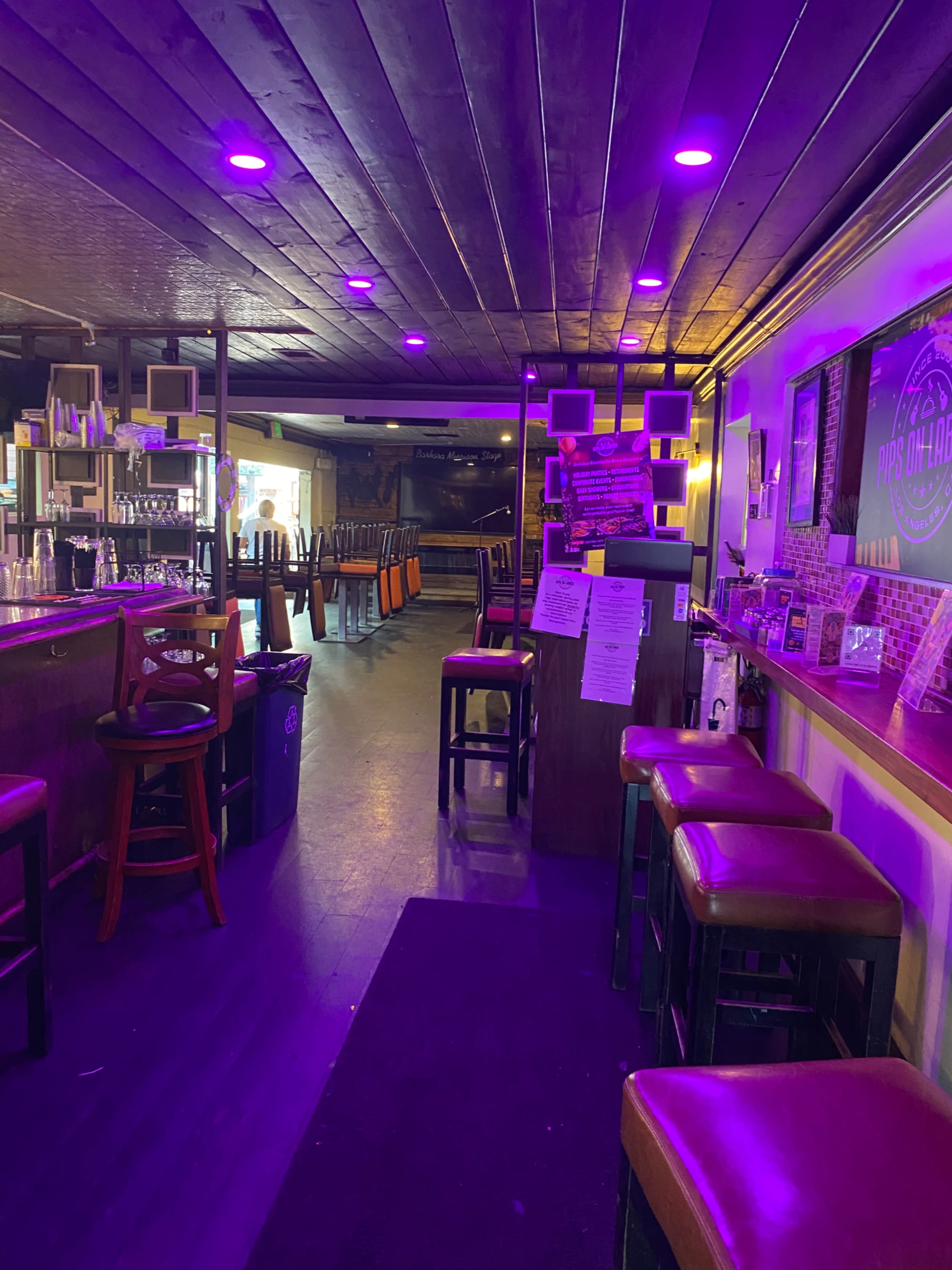 The image shows the interior of a bar with dark wooden flooring, purple lighting, and empty high-top tables and chairs arranged across the space.