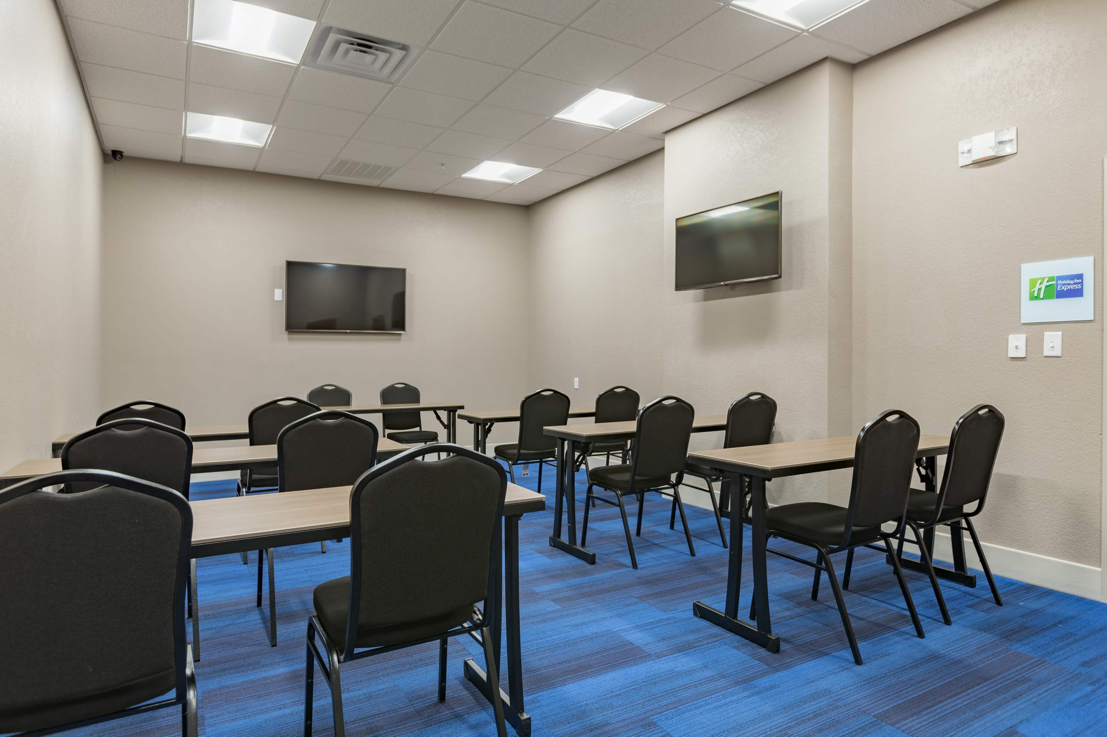 A meeting room with multiple rectangular tables and black chairs, along with two wall-mounted televisions.