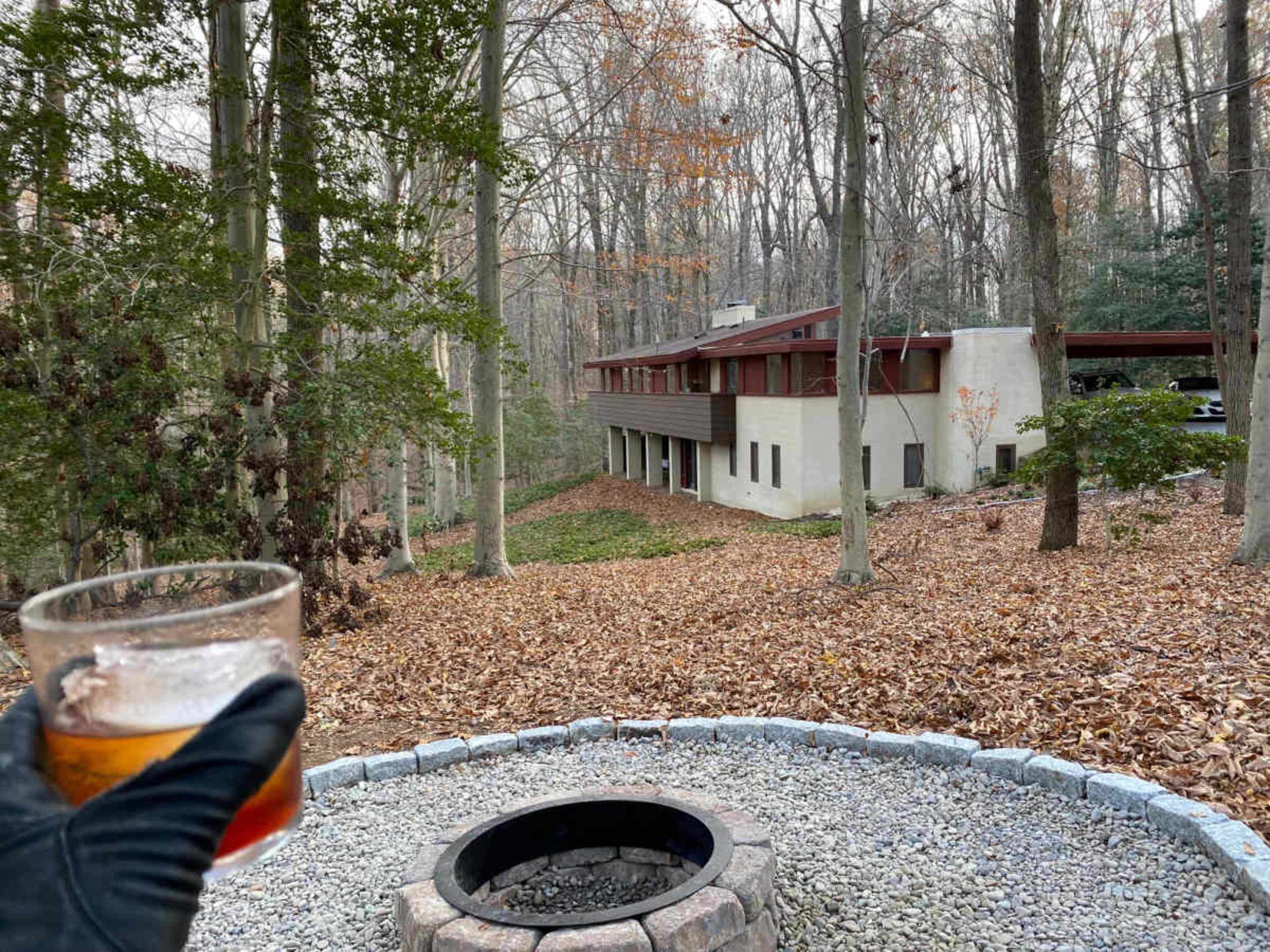 A person holding a drink stands in a wooded area with a view of a modern house and a stone fire pit surrounded by fallen leaves.
