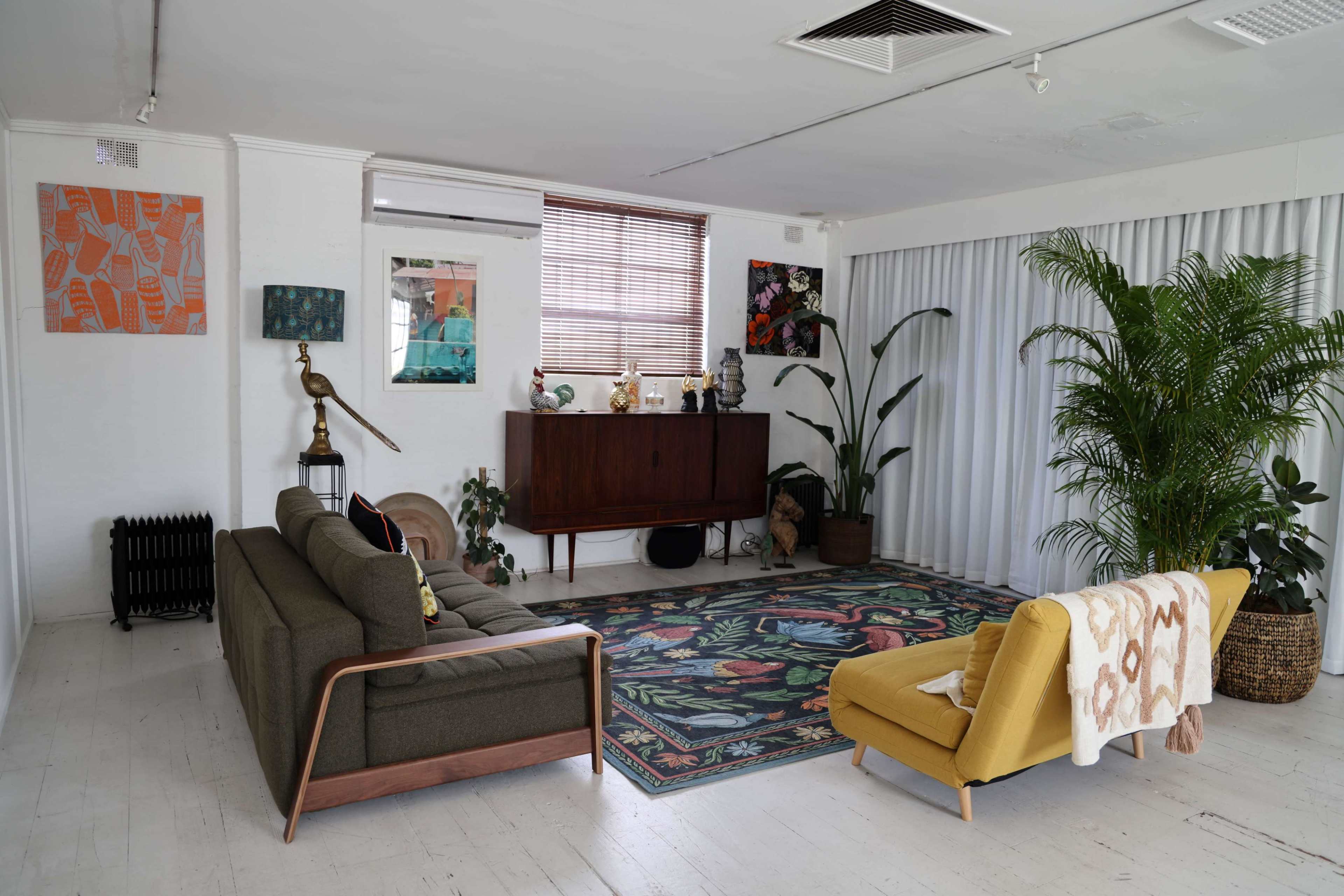 A spacious living room features a green sofa, a yellow chair, a decorative rug, indoor plants, and art on the walls.