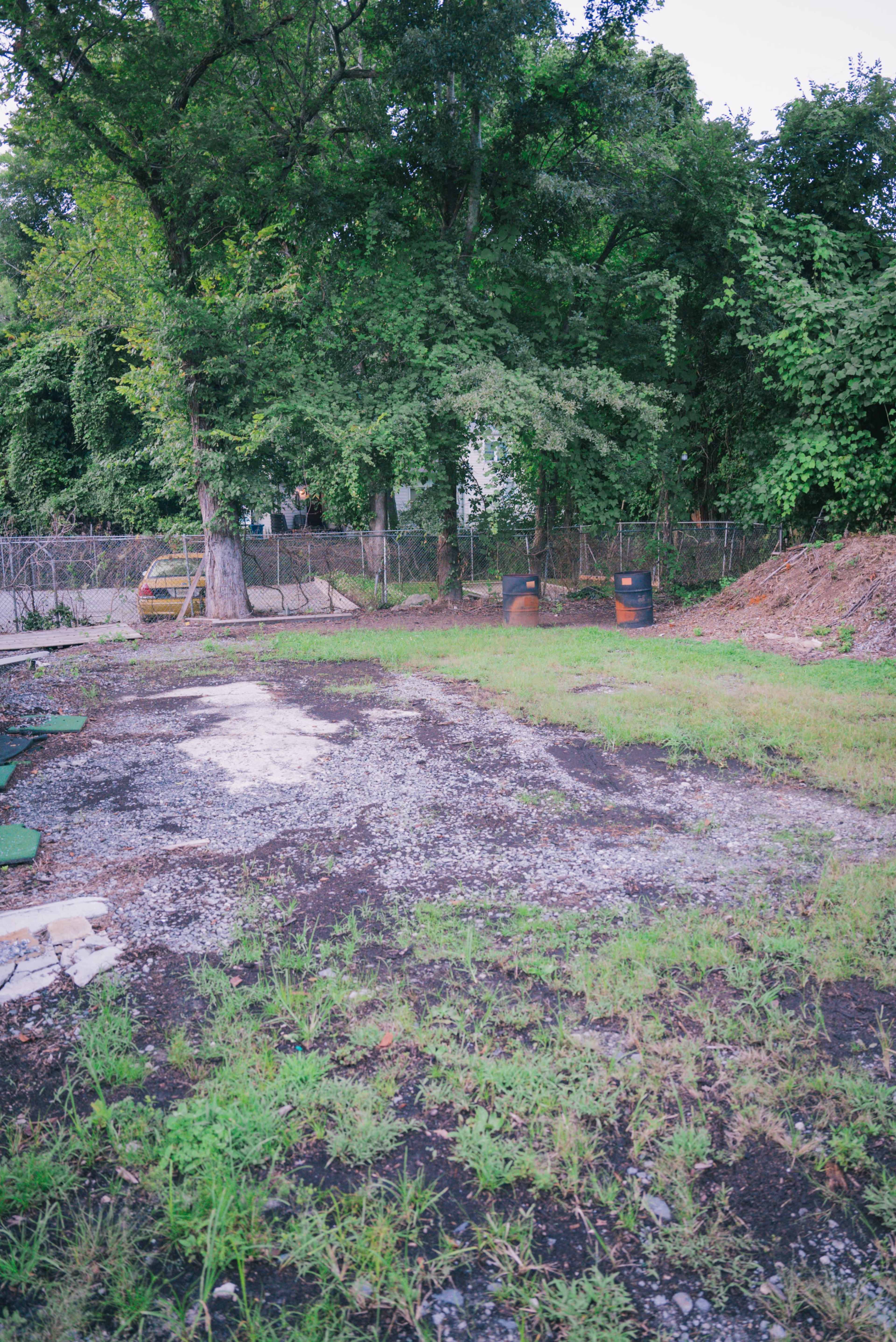 The image shows an overgrown, gravel-covered lot surrounded by trees and a fence, with two barrels visible in the background.