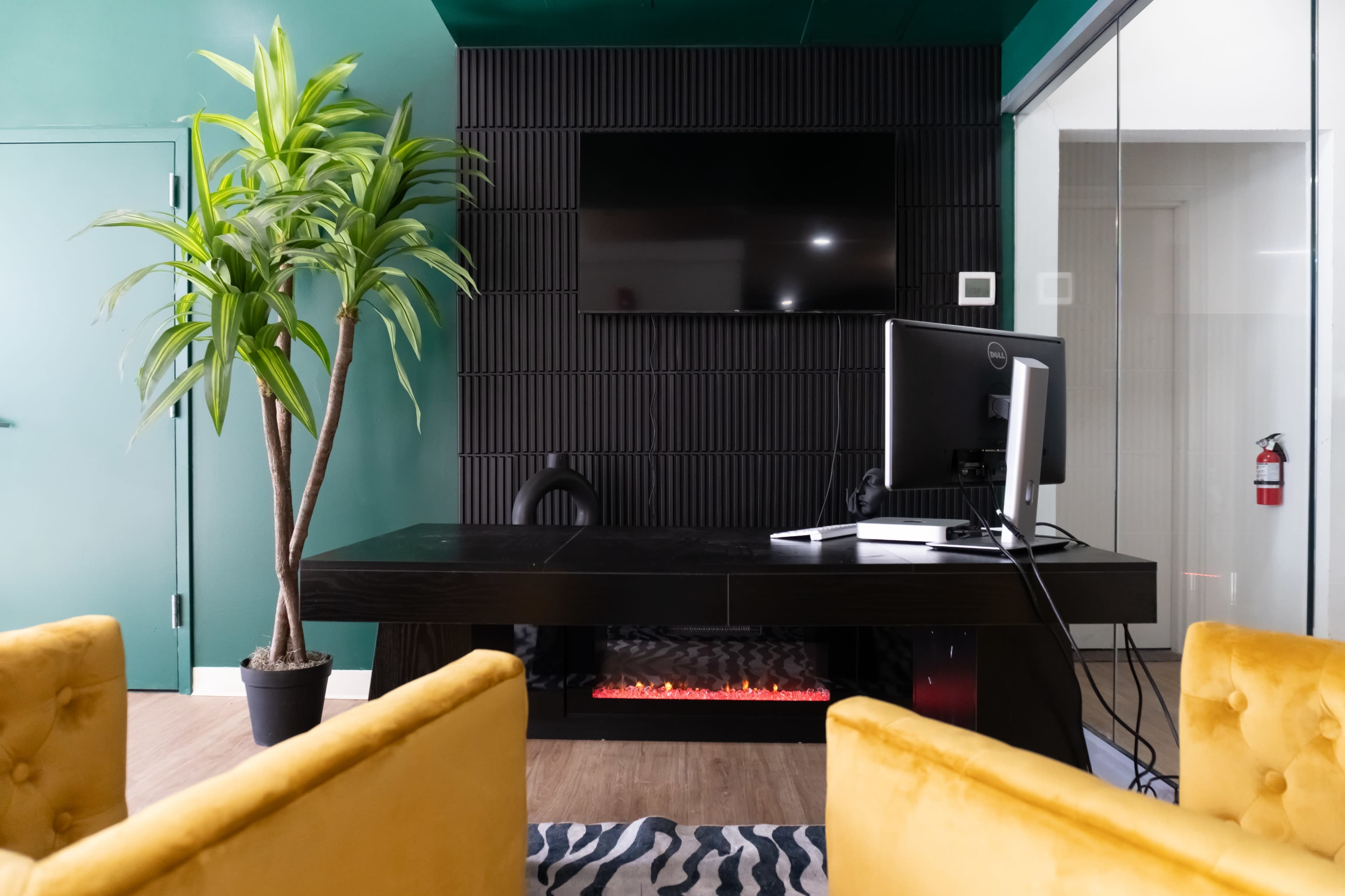 The image shows a modern office space with a dark wall-mounted television, a potted plant, a desk with a computer, and two yellow armchairs facing a faux fireplace.