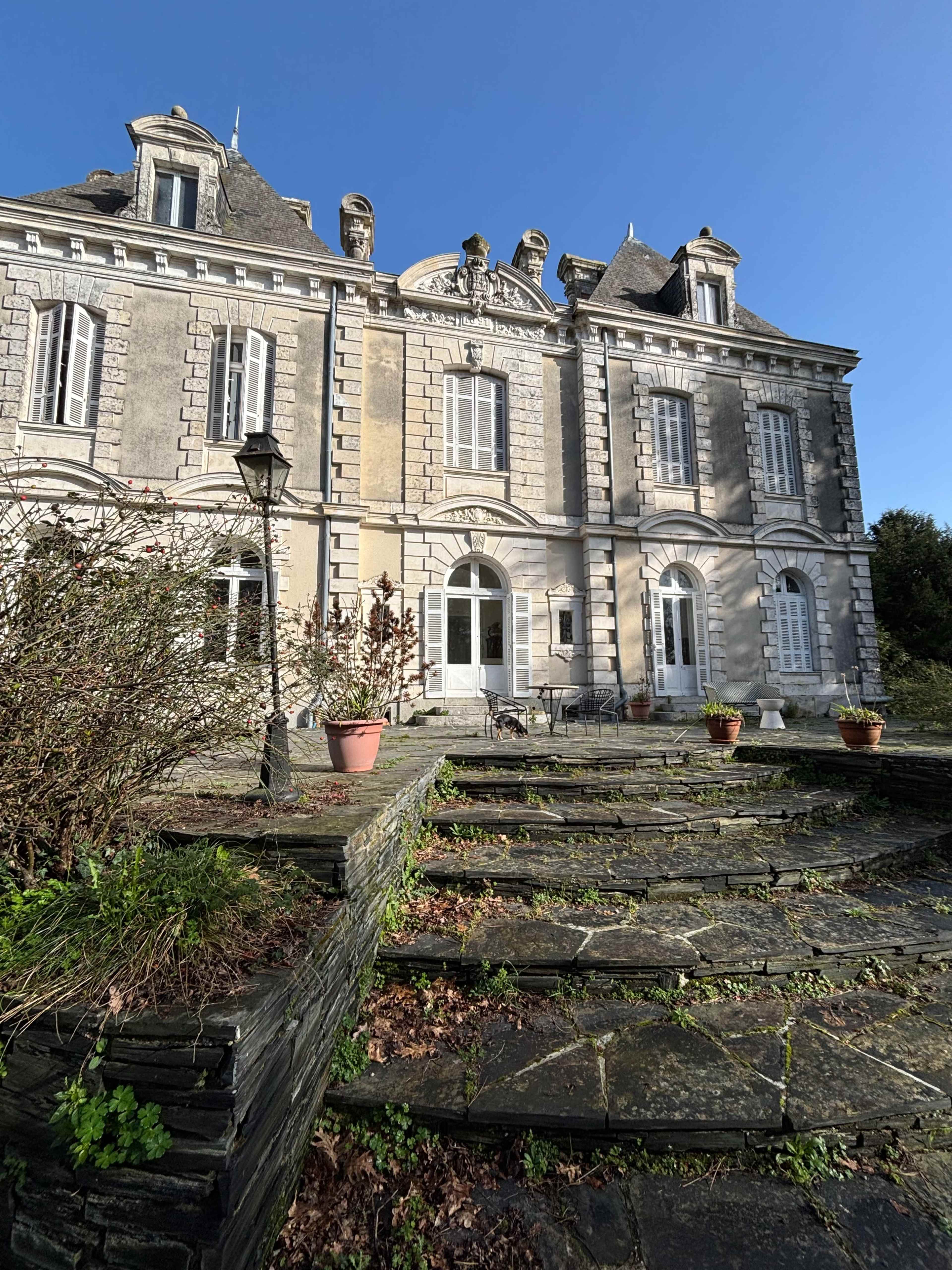 A large, historic stone mansion with a symmetrical façade, surrounded by neatly trimmed gardens and a stone staircase leading to the entrance.