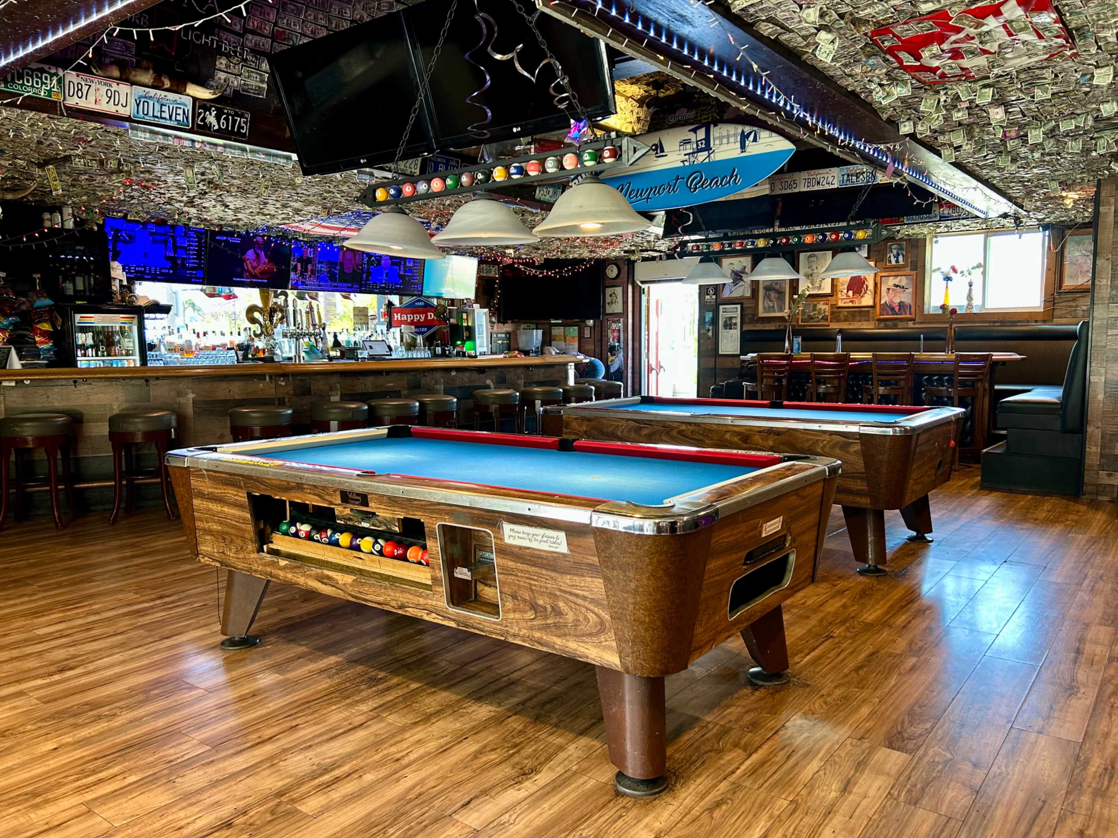 The image shows a bar area with two pool tables, wooden flooring, and a ceiling decorated with various items, including photographs and license plates.