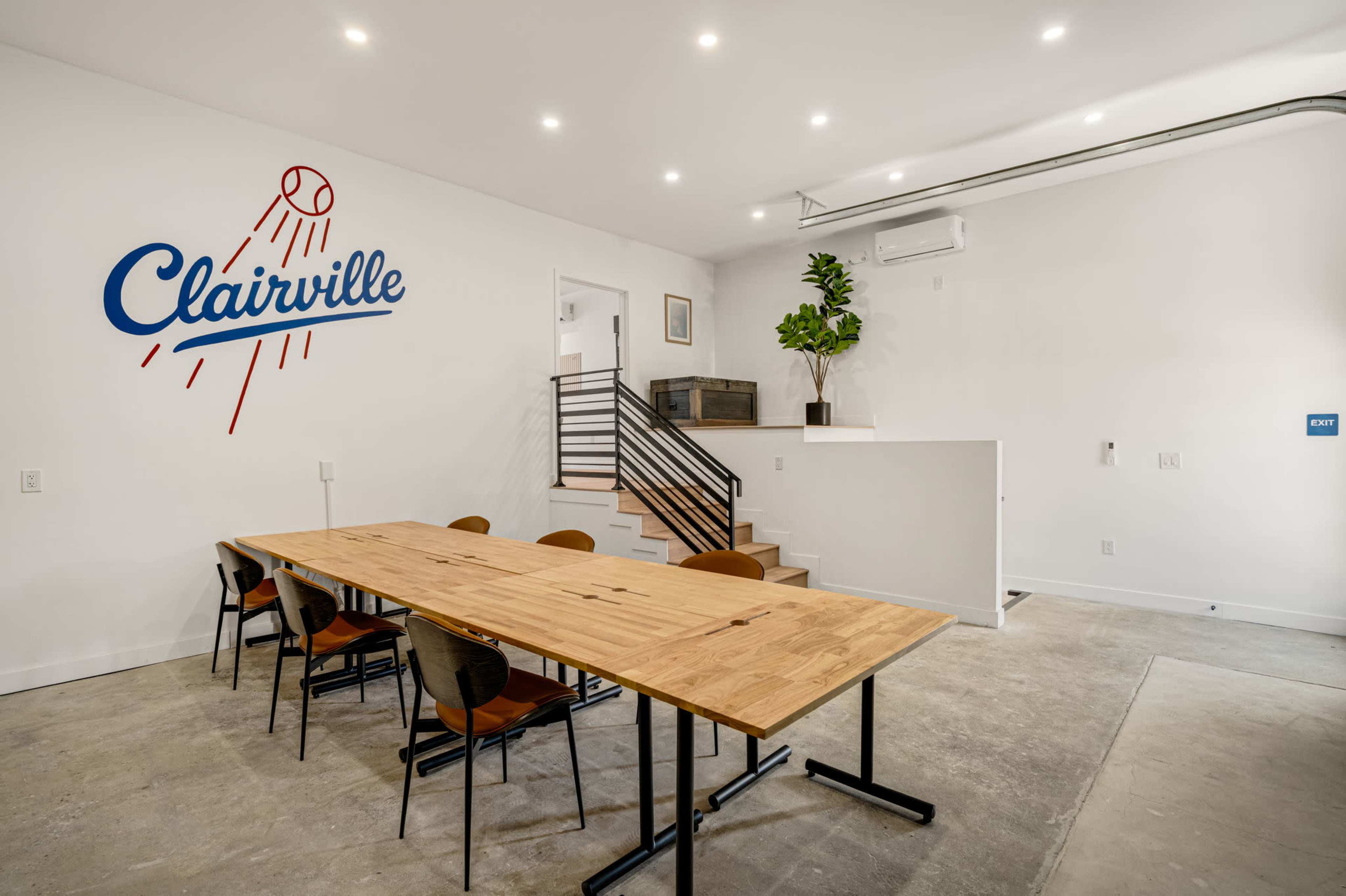 The image shows a modern meeting space featuring a large wooden table with several chairs, a minimalist design, and a wall adorned with a baseball-themed logo reading "Clairville."