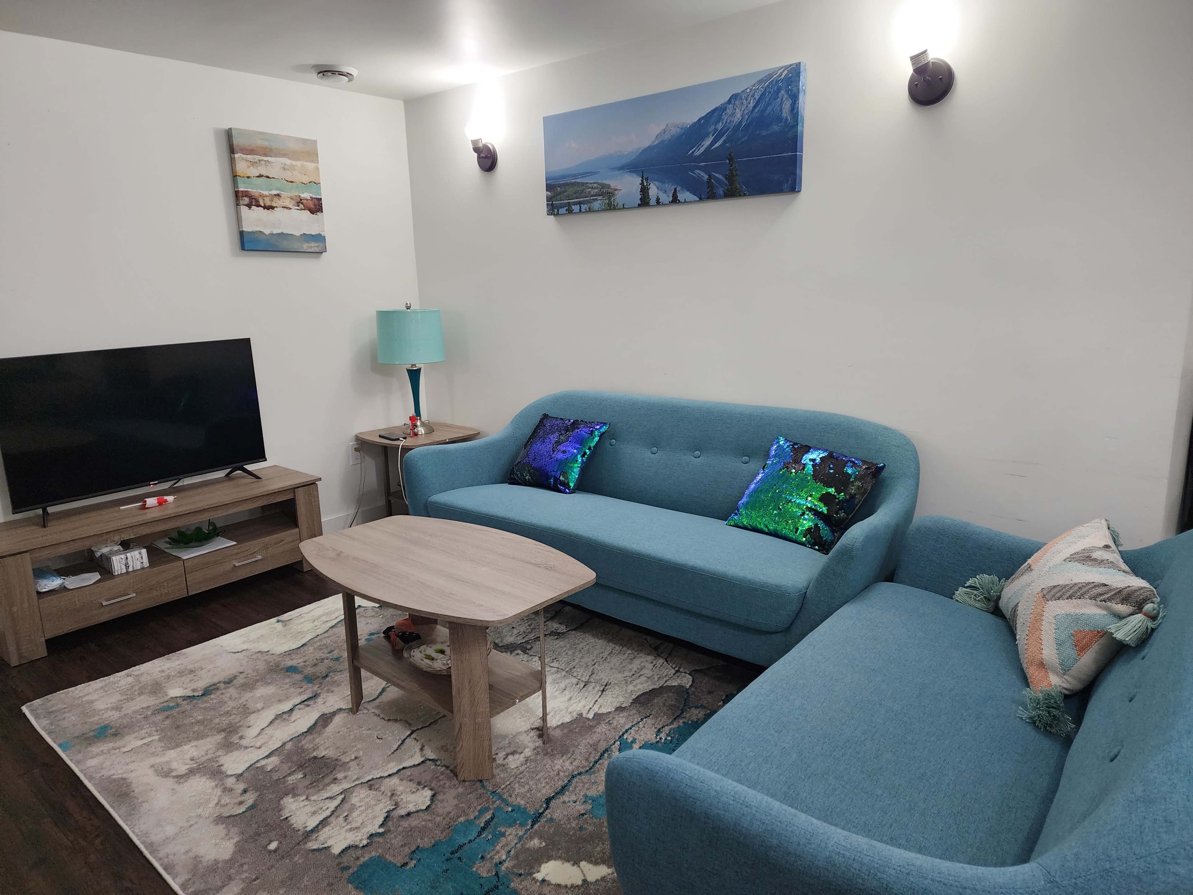 The image shows a modern living room featuring a blue couch, a matching armchair, a coffee table, a television on a media console, and decorative wall art.