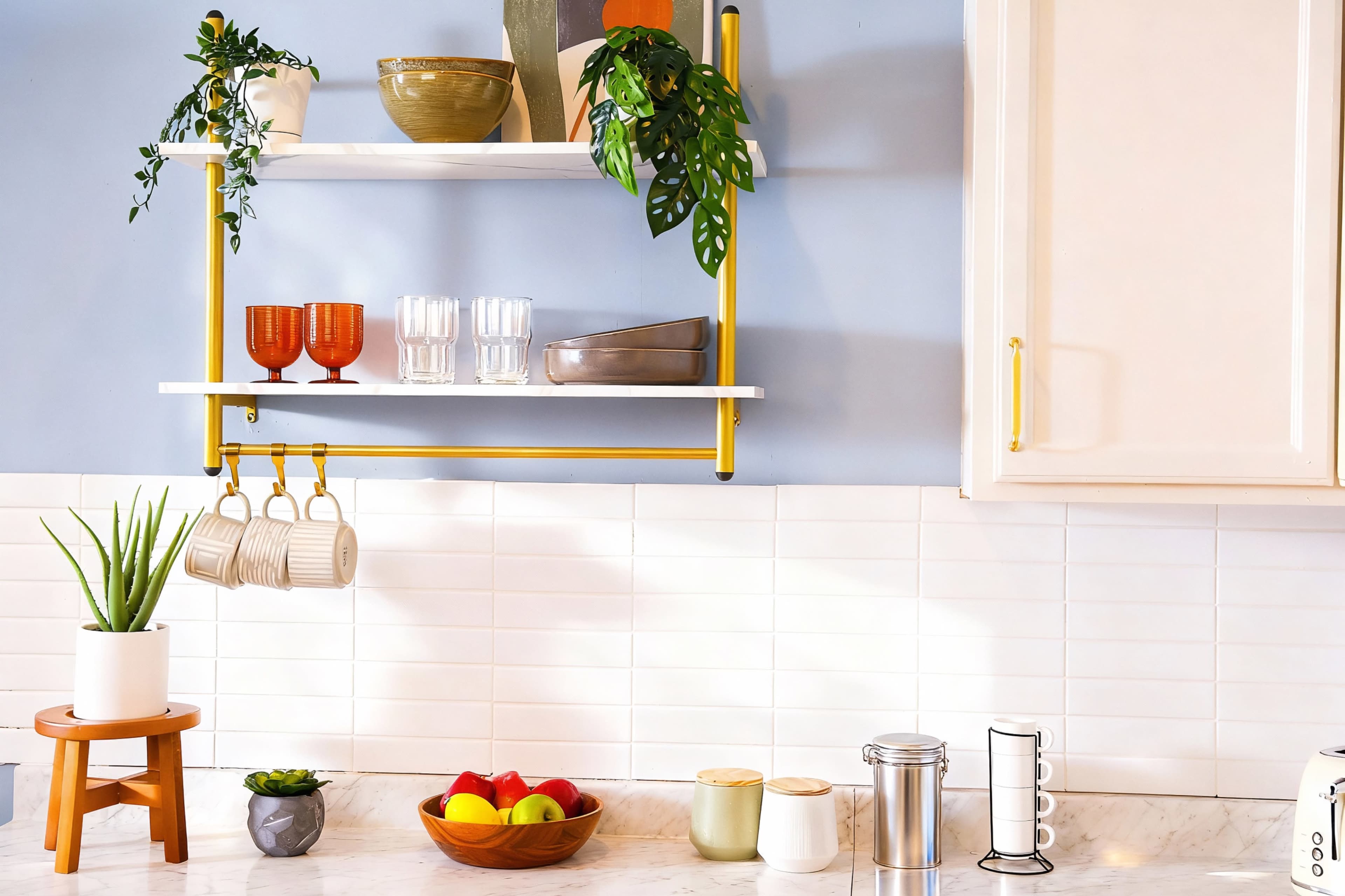 The image shows a modern kitchen shelf displaying various dishes and plants above a marble countertop with fruits and a few kitchen accessories arranged neatly.